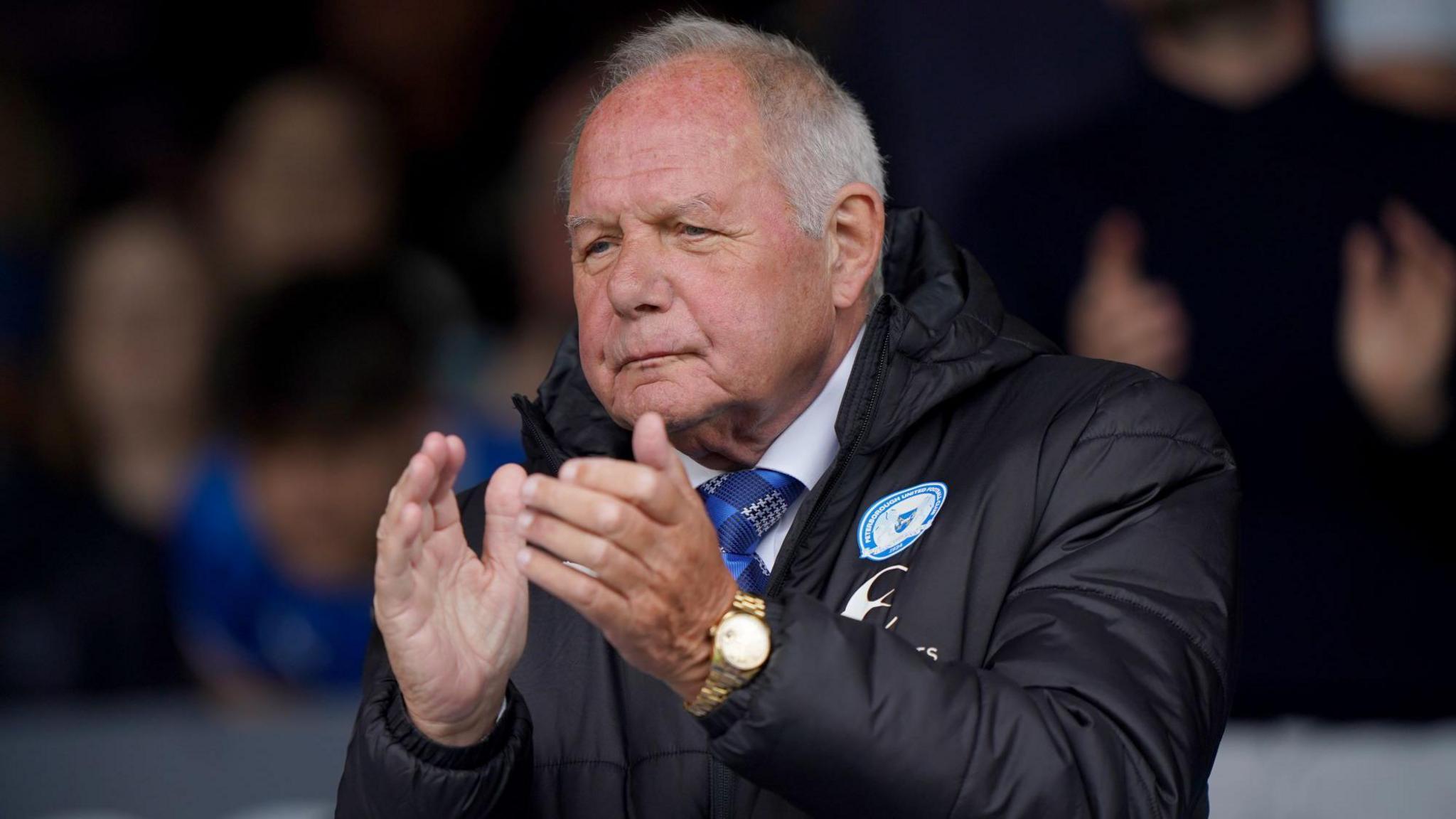 Barry Fry clapping his hands while wearing a black Peterborough United coat. Fry has short grey hair and also has a blue tie on and a gold watch.