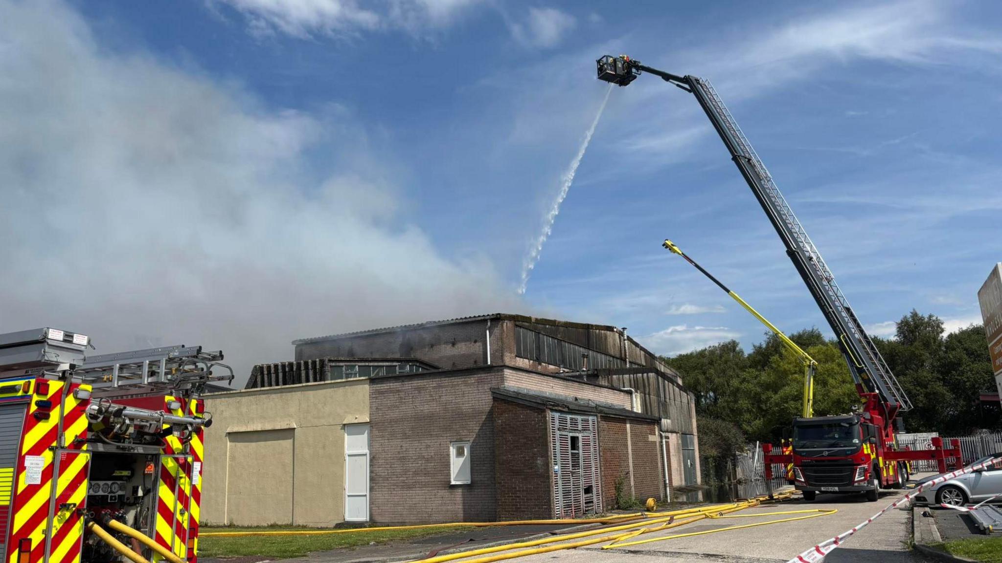 Water is hosed down onto a one-storey building from a firefighter on a cherry picker. Fire engines can be seen around the building