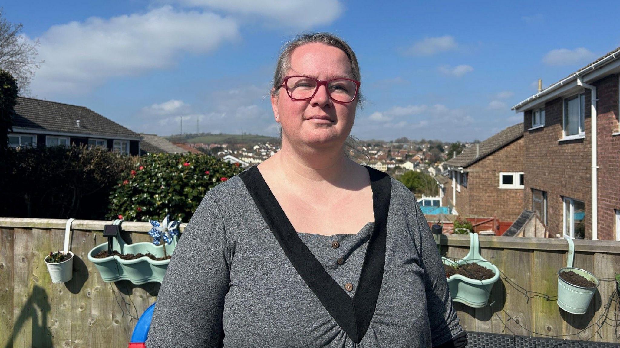 A woman wearing a grey top and pink glasses standing in her garden with flower pots behind her