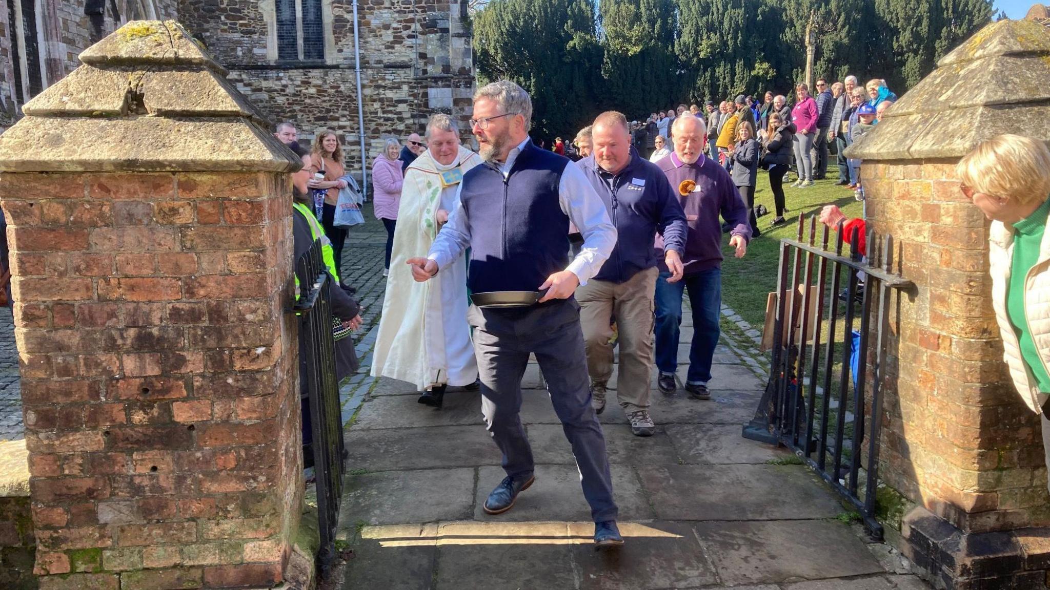 A group of men holding frying pans run out of Wimborne Minster church. There are crowds of people watching, with the church in the background.