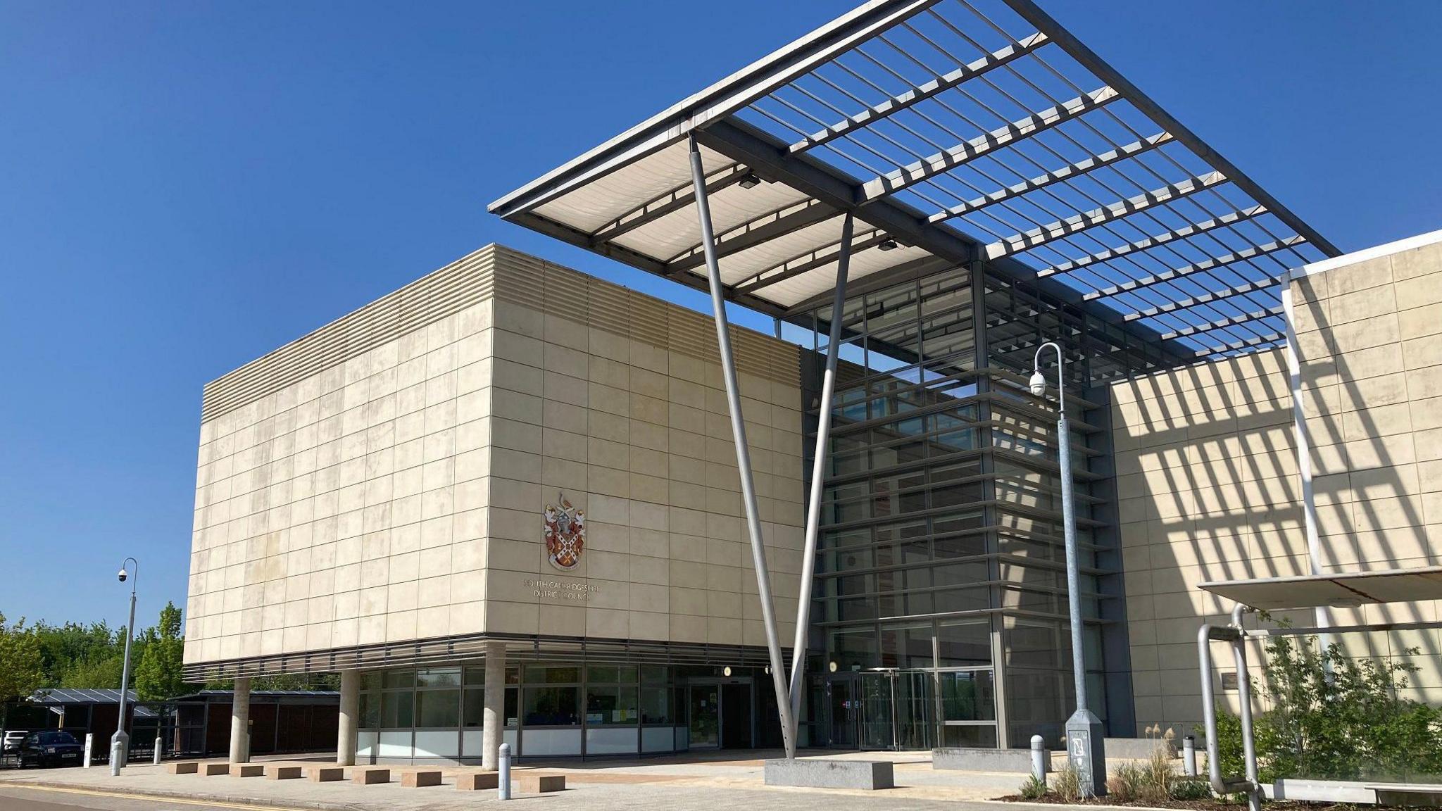 South Cambs council building which is a modern blocky structure made of beige blocks and a recessed glass section in the middle. There is a large dark grey metal pergola structure held up by V-shaped poles. Clear blue sky behind.