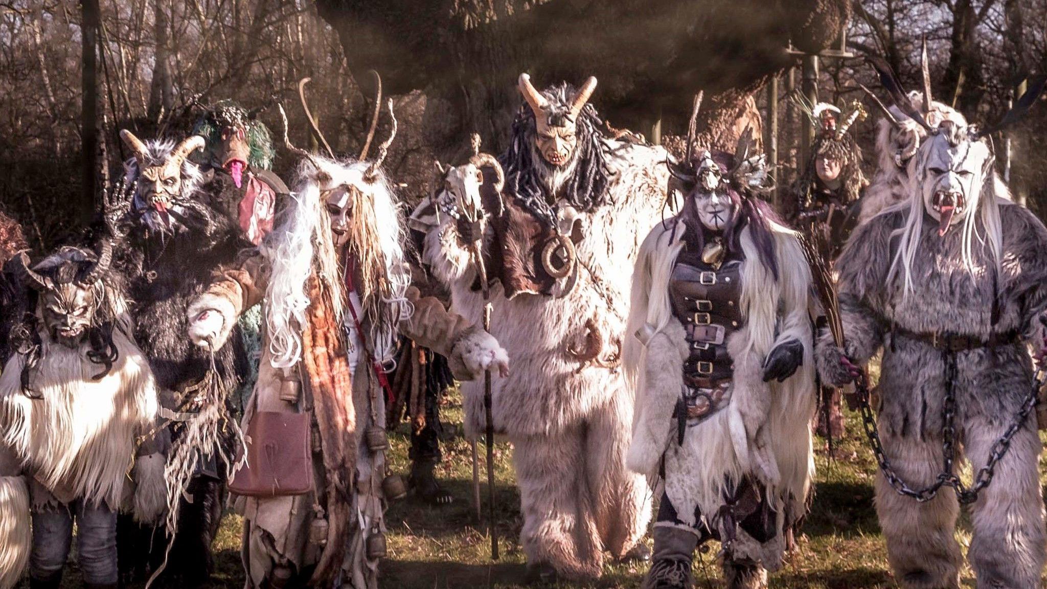 The Krampus wear elaborate, demonic-inspired animal costumes. Here they are at a previous event walking through a sun-dappled forest, creating an eerie scene. There are six of them in white in this image.