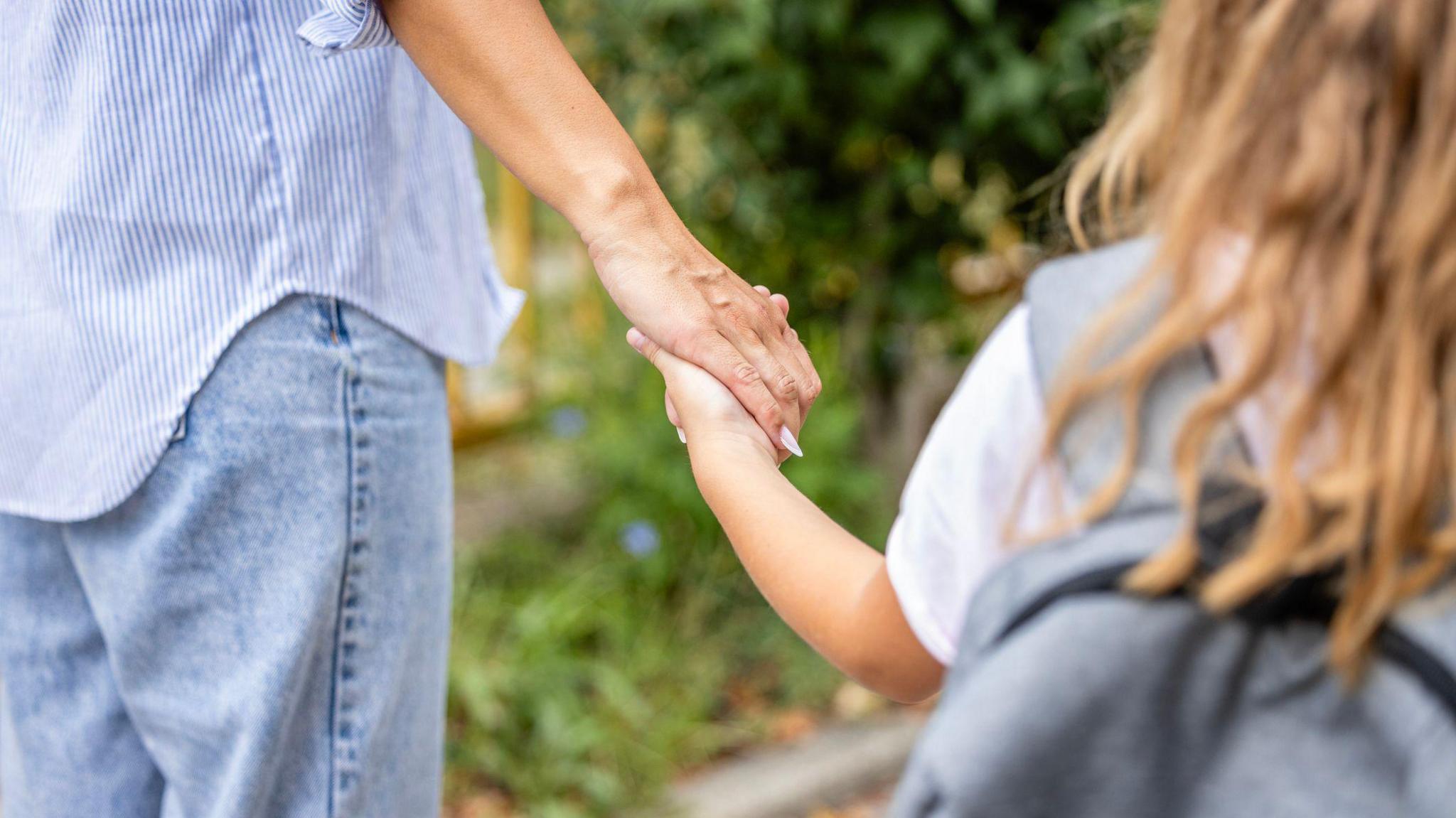 A woman in blue jeans and a blue and white striped shirt is holding hands with a young girl who has brown hair and a grey backpack. Only the backs of them are visible.