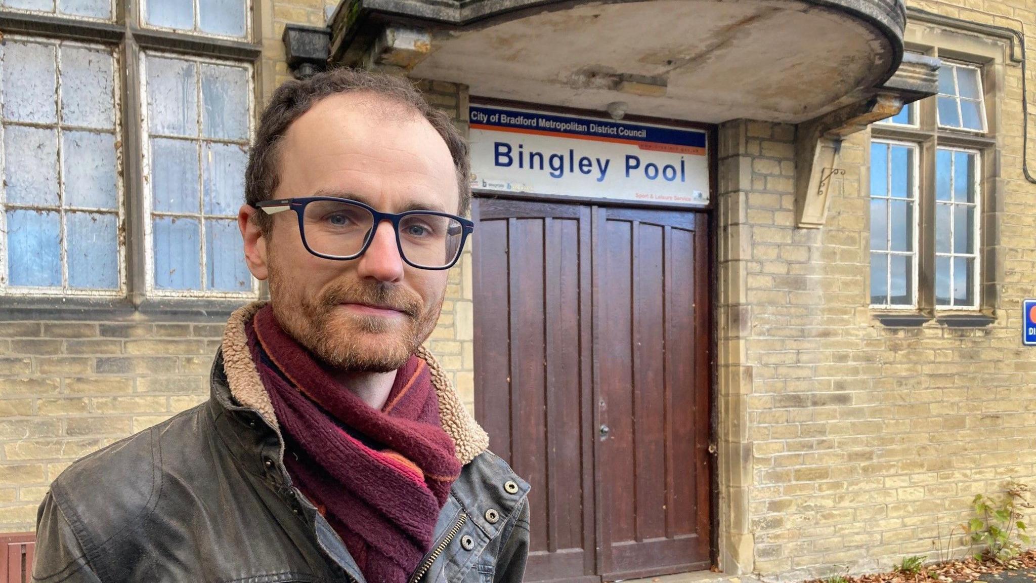 A man in glasses standing in front of two wooden doors with a white sign above which reads Bingley Pool in purple lettering