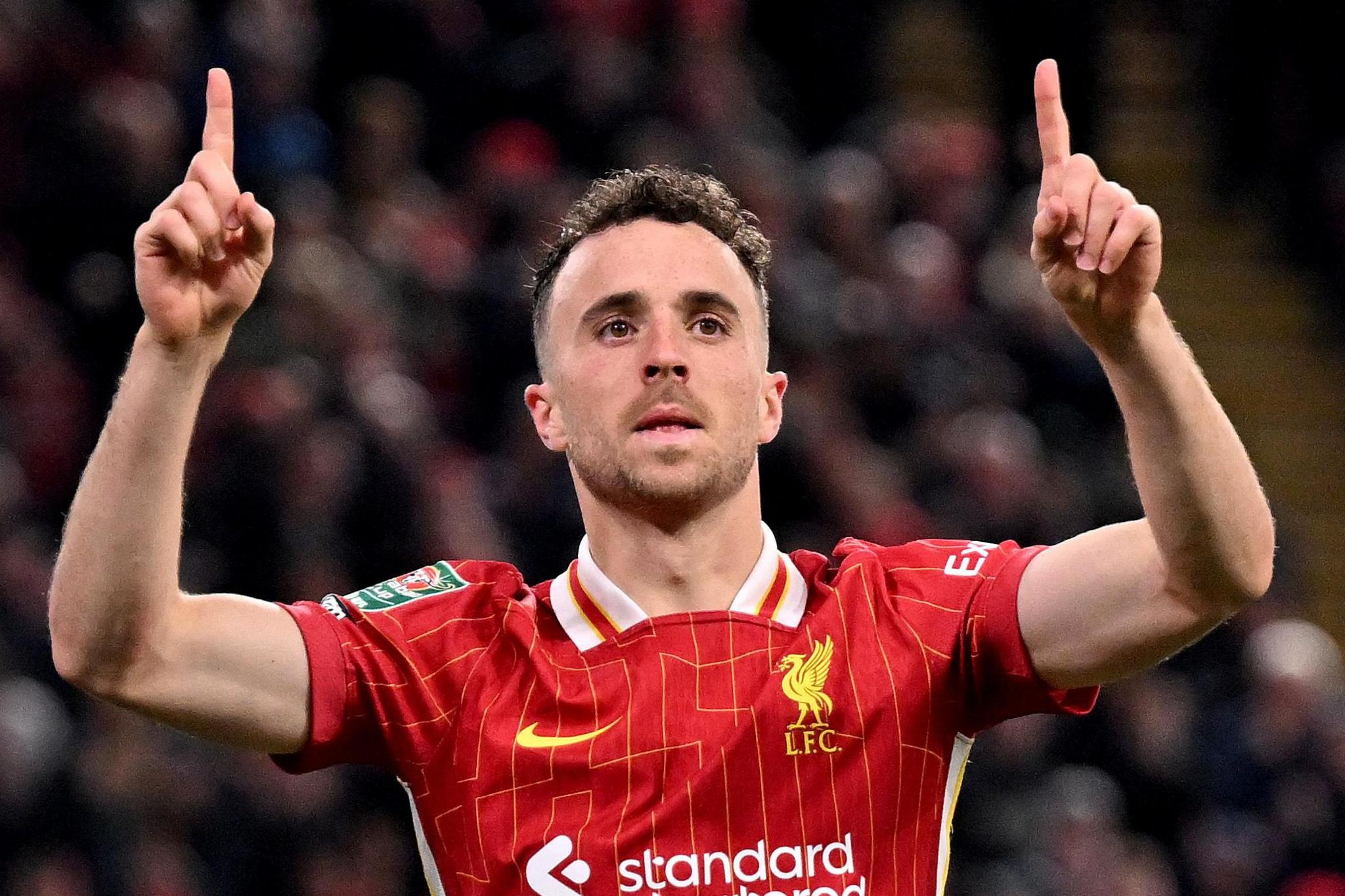 Diogo Jota, i veshur me një fanellë të kuqe të Liverpool FC me vija të verdha dhe stemën e klubit, ka ngritur të dy krahët me gishtat tregues që tregojnë lart në një gjest festiv. Fanella ka logon e sponsorit "standard chartered" dhe një detaj me jakë të bardhë. Sfondi tregon një turmë të turbullt në një stadium.