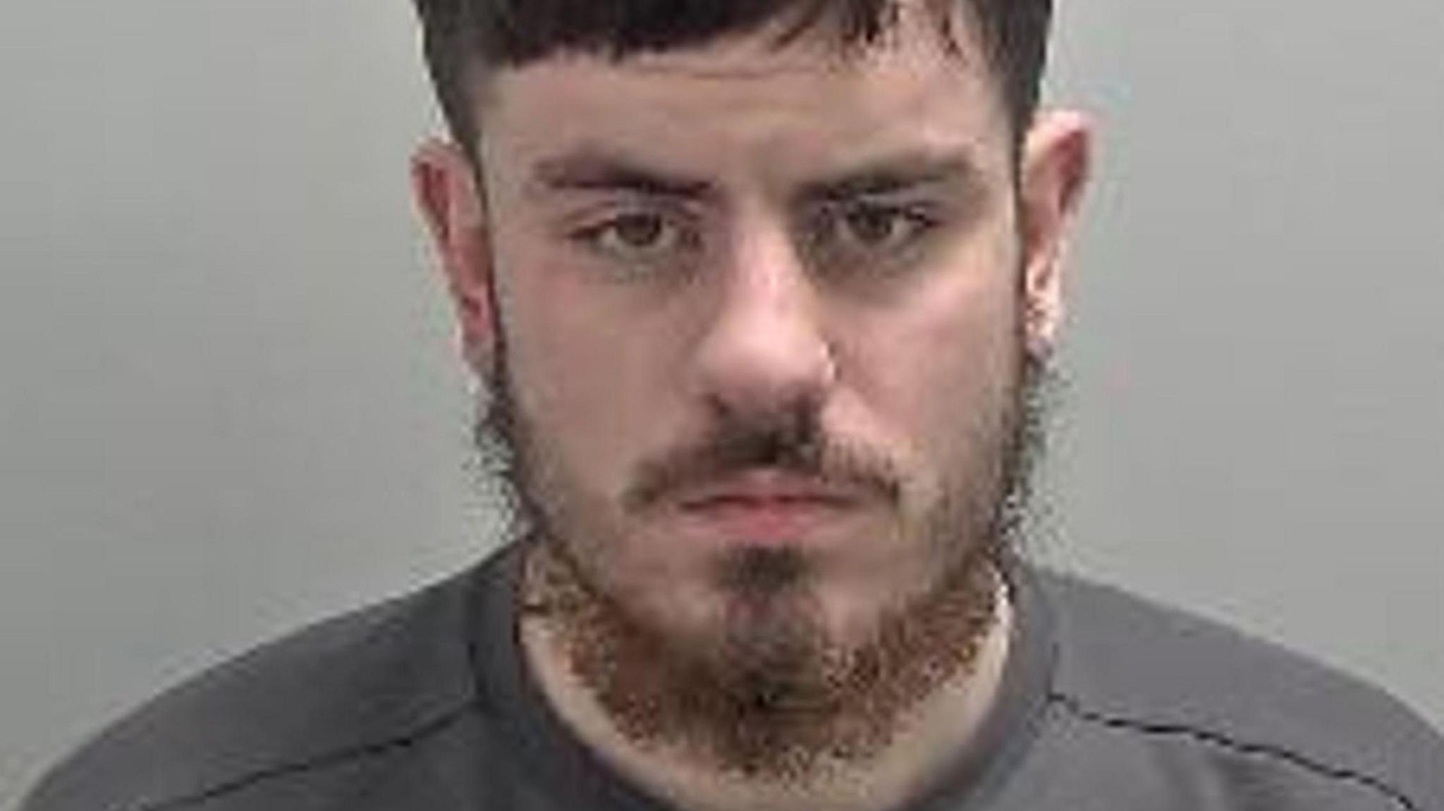 Frankie Maughan in his mugshot photo. It is low quality, he has a beard and is stood against a plain background. 