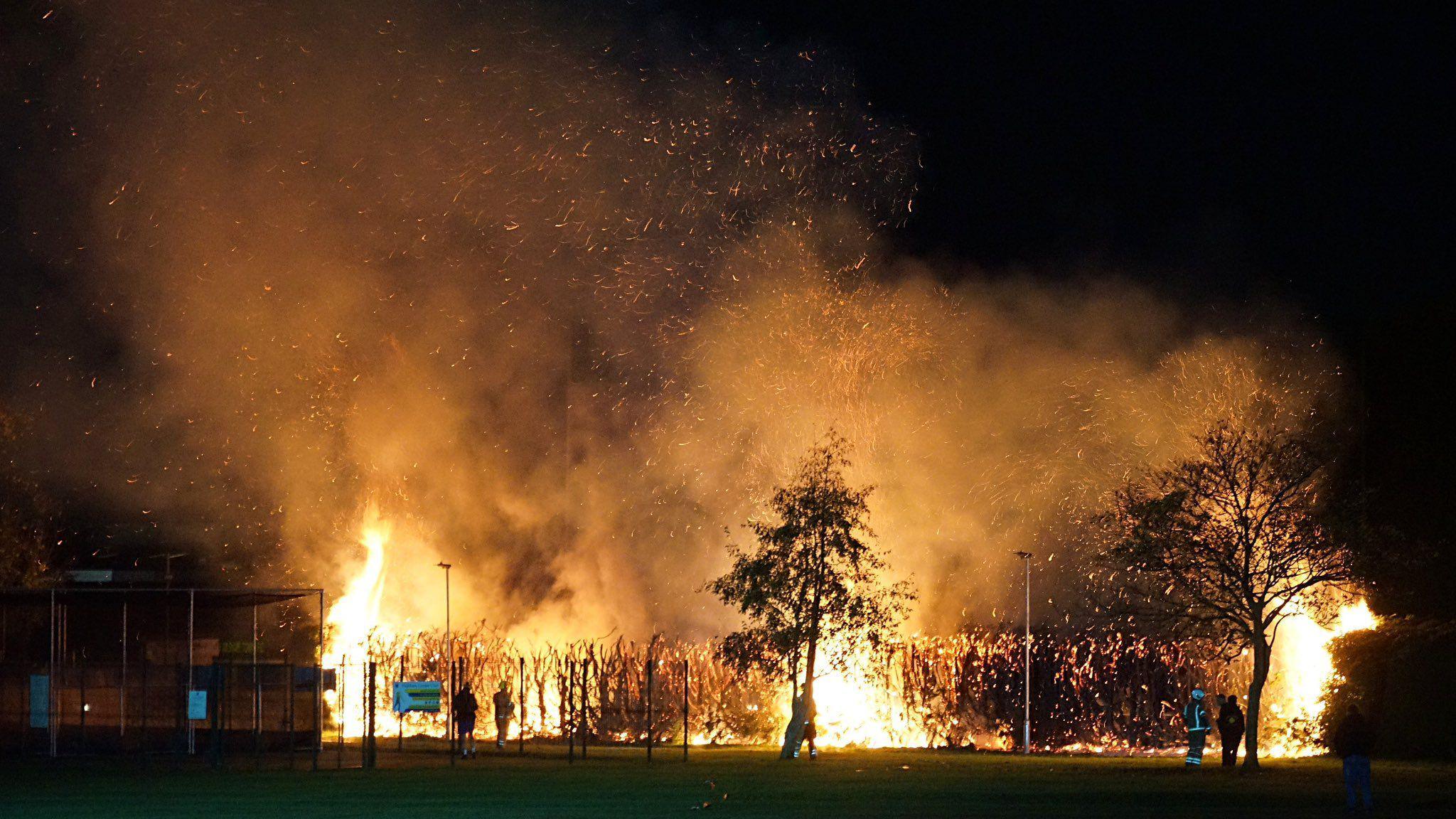 A fire burns at night and large flames create a silhouette of trees and a hedge, and a football stand is seen to the left of the image. Sparks are flying in to the air.