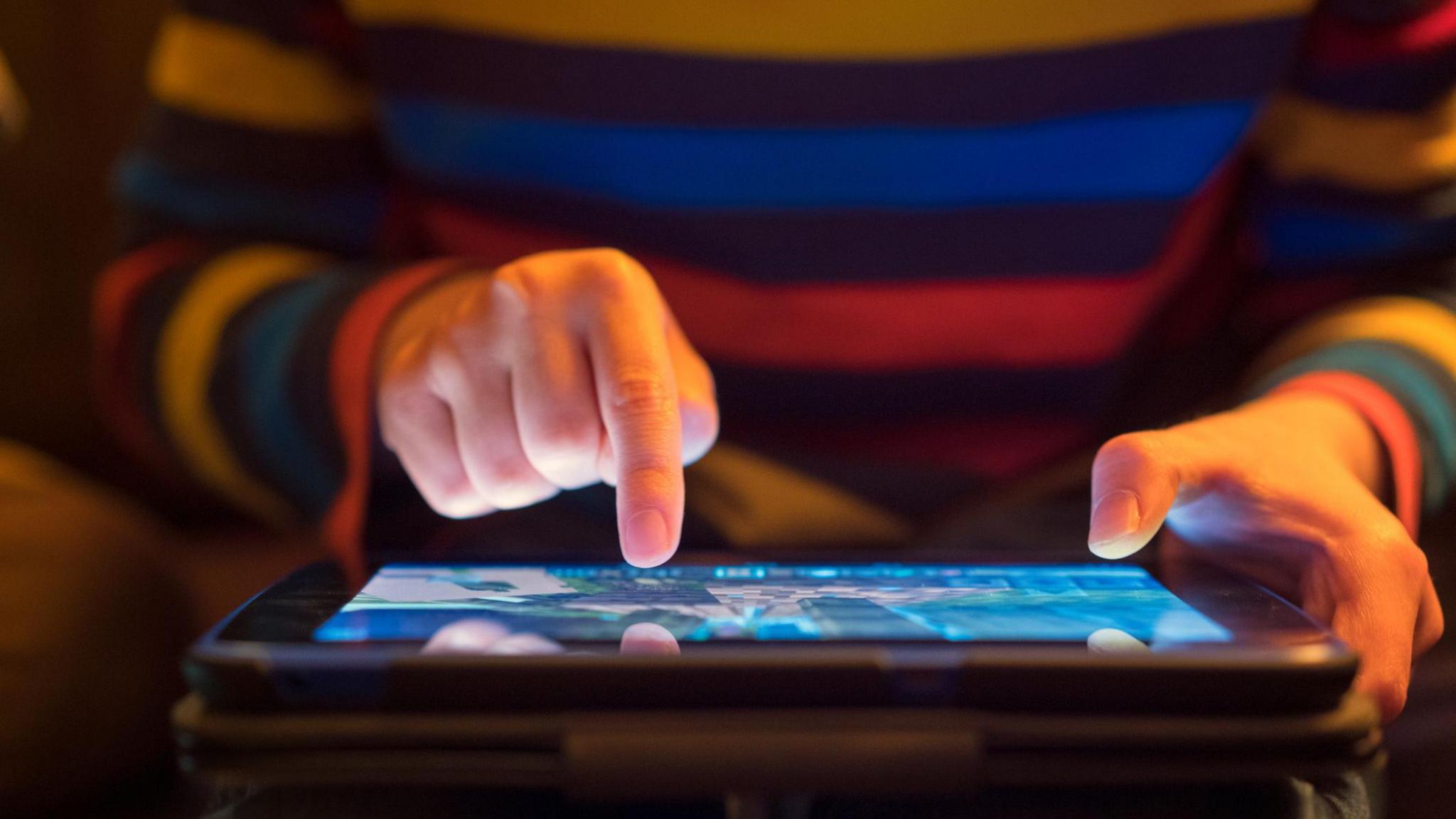 An image of a person using an iPad. It shows a child holding the device with two hands, one holding the iPad and another with one finger on the screen. The child is wearing a colourful stripy jumper