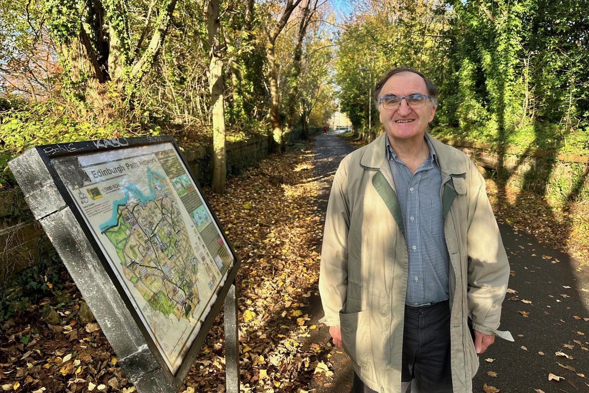 Lawrence Marshall is standing on the Roseburn Path beside a board showing all the defunct rail routes in Edinburgh. The path is lined with trees and there are yellow and brown leaves on the ground. He is wearing glasses and a long, green jacket and has brown receding hair. He is smiling at the camera.