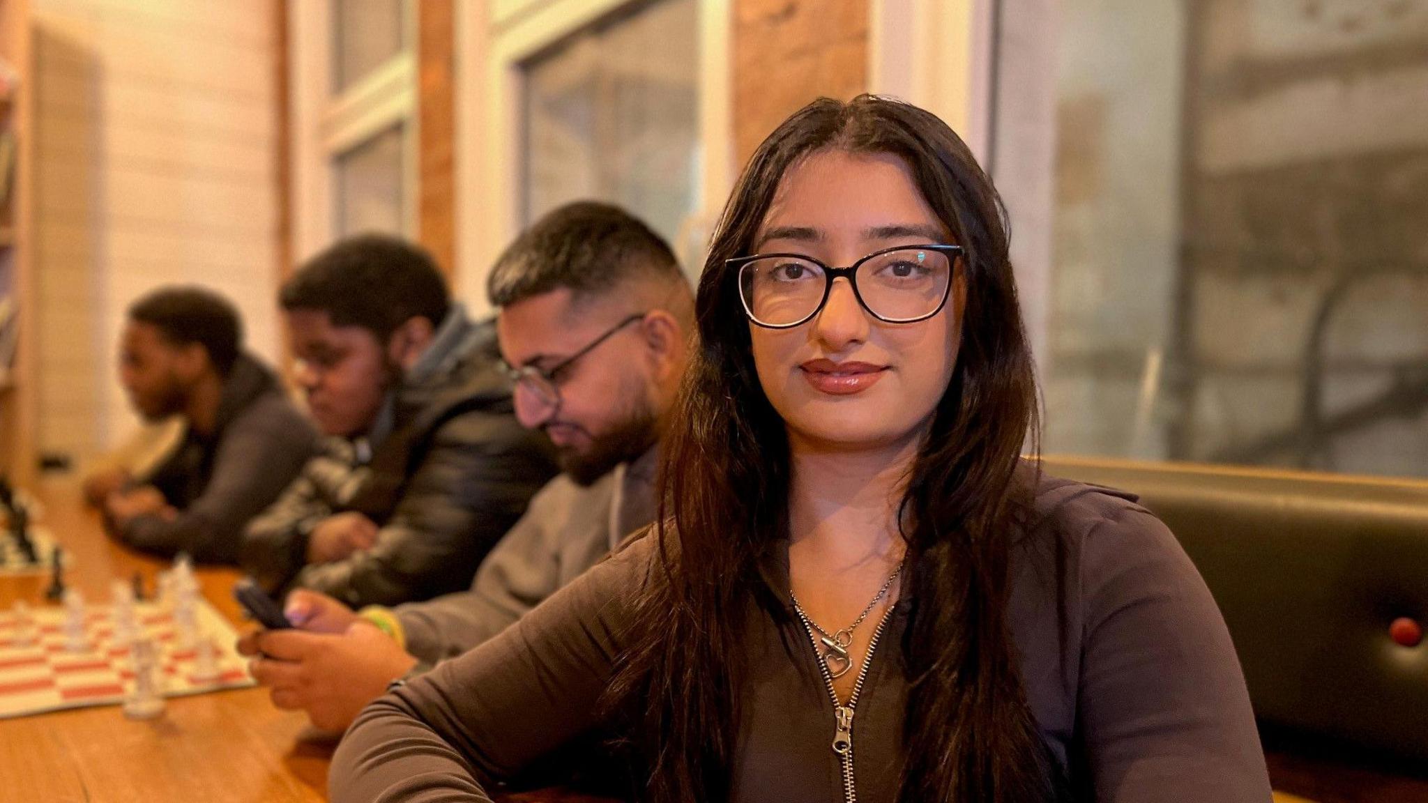 A young woman with long brown hair and wearing glasses looks at the camera. she has a brown zipped up top on. Three men sit behind her, one looking at his phone, the other two at chess boards.