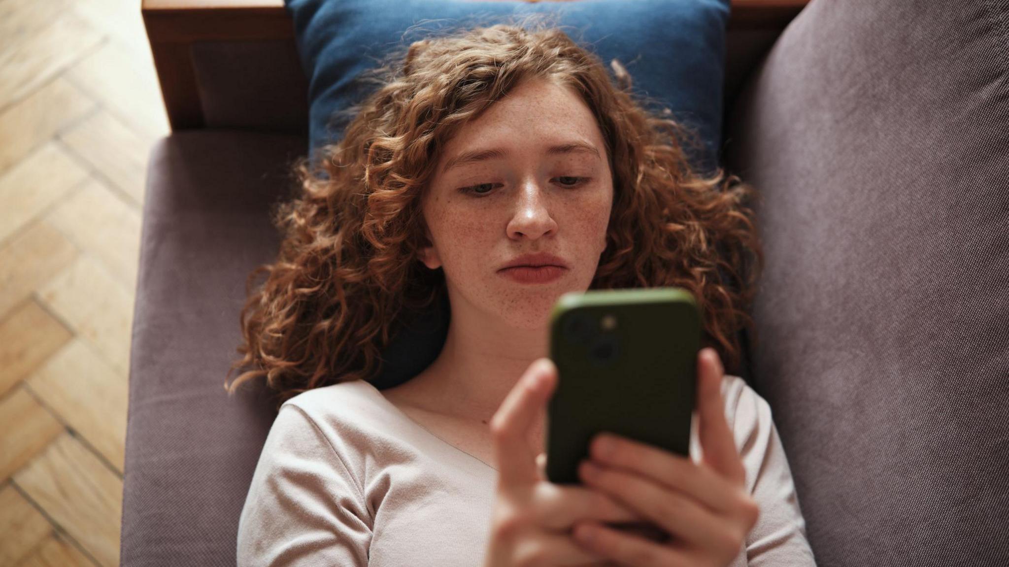  A teenager lies on a grey sofa looking at her phone, which has a green case. She has curly red hair and freckles, and wears a cream top.