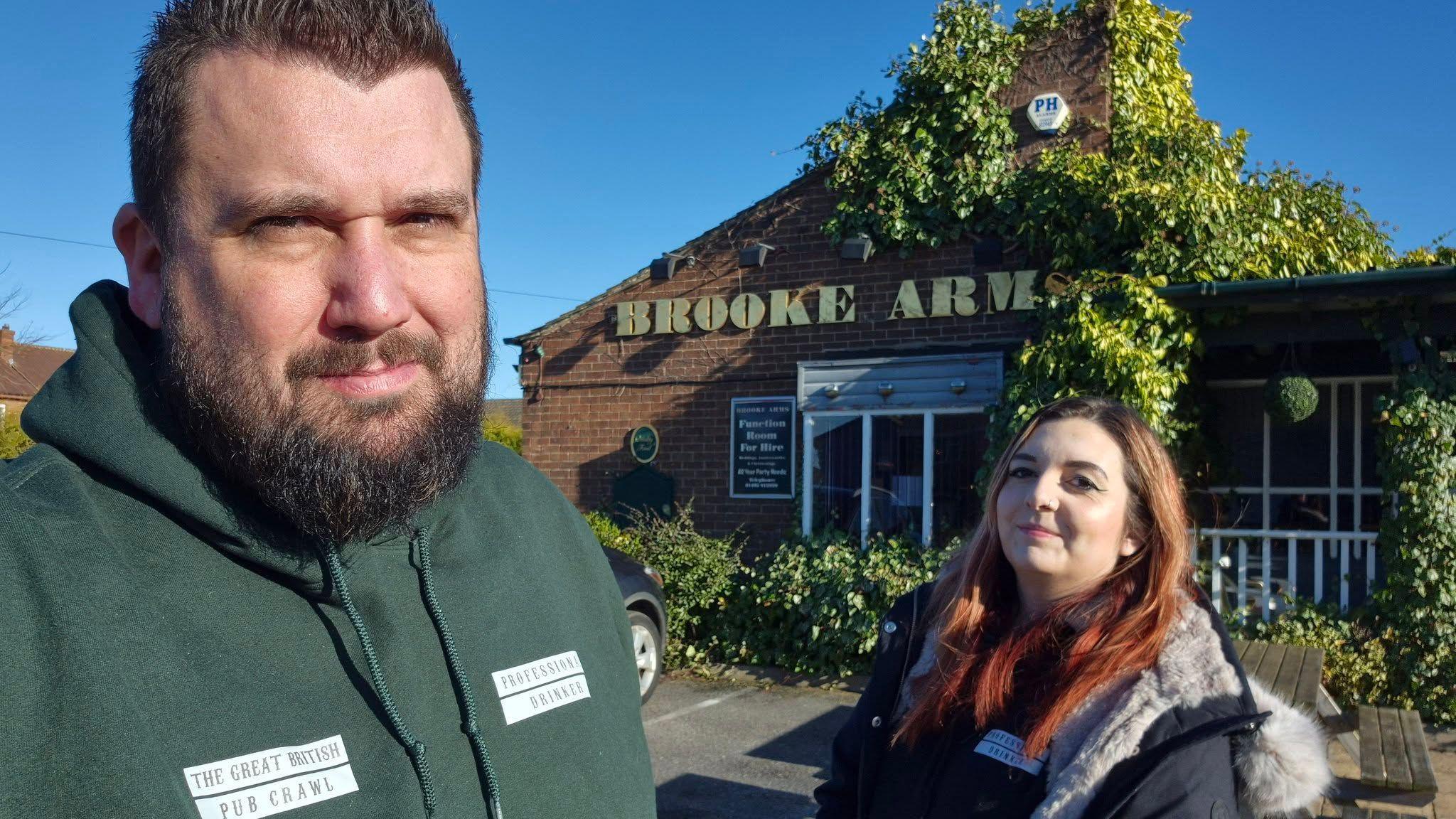 Full outdoor shot of a man and a woman standing in front of pub called the Brooke Arms, which is covered in Ivy. 

