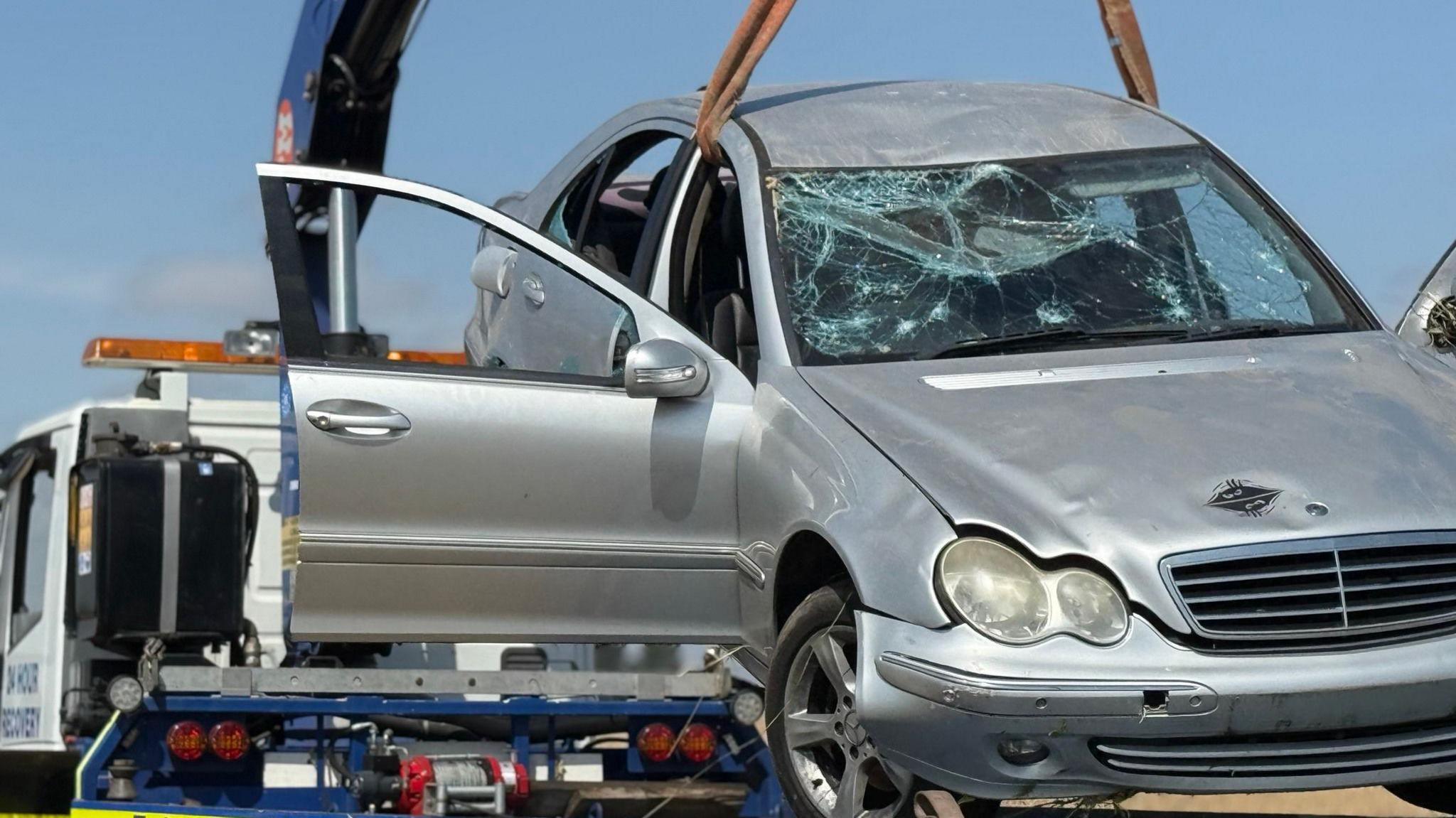 A large silver car is being winched on to a recovery truck. The car has a smashed front windscreen and both the driver and passenger doors are open