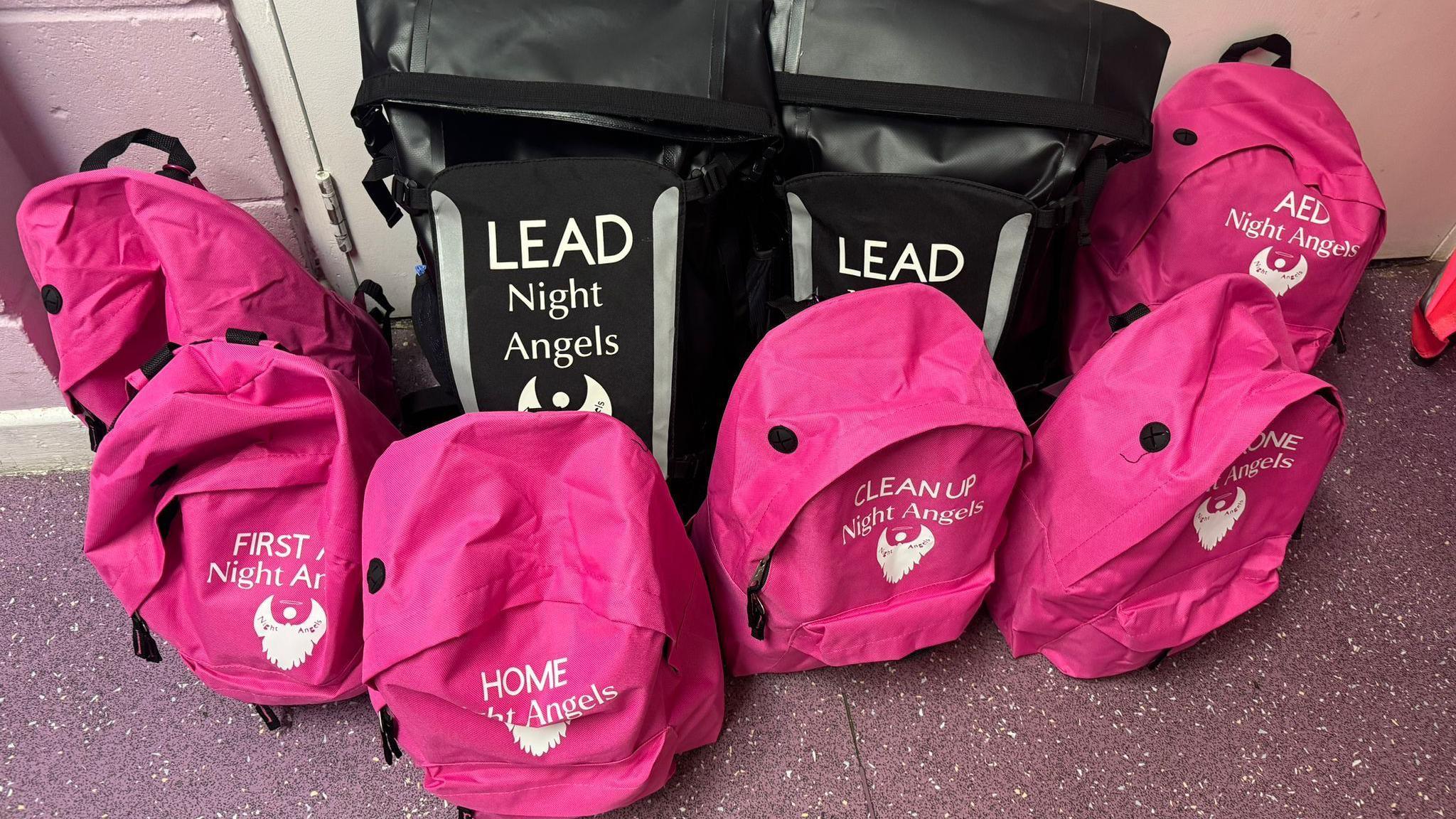 A pile of pink backpacks printed with the Night Angels logo in white. Each one is labelled to match their respective roles and purposes, including first aid, AED and clean up. Two larger black backpacks are also among the pile - they are also printed with the Night Angels logo and labelled with the word lead in capital letters. 