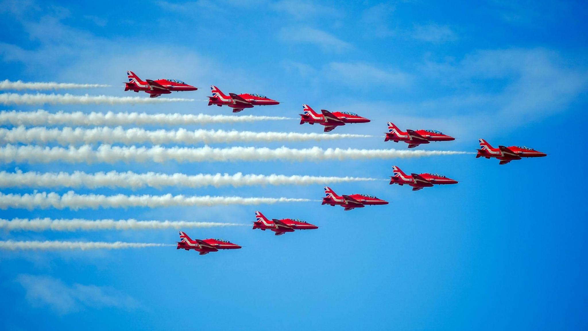 Nine red planes fly in formation through the sky. A trail of white smoke follows each of them. The aircraft are flying in an arrow shape against the backdrop of a brilliant blue sky.