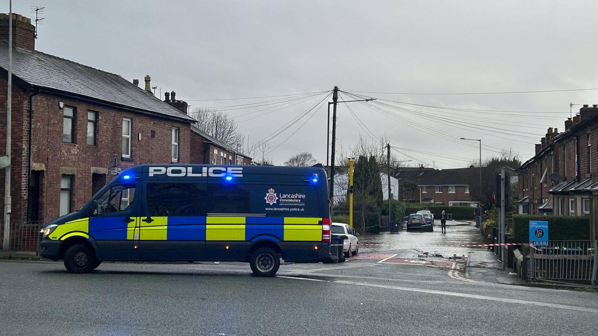 A large dark blue Lancashire Police van is parked at the entrance to a street lined with houses, which is partially submerged