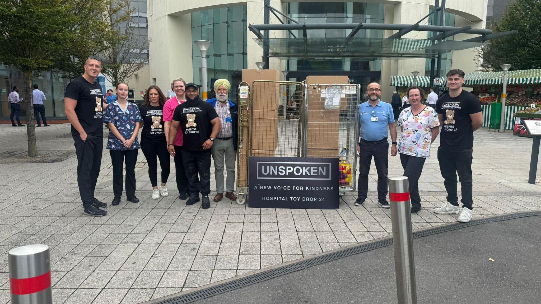 Atiq and his team of volunteers and some hospital staff members pose outside a hospital with boxes of toys. At the front of the boxes a sign is propped up which reads "Unspoken: A New Voice For Kindness Hospital Toy Drop 24".  