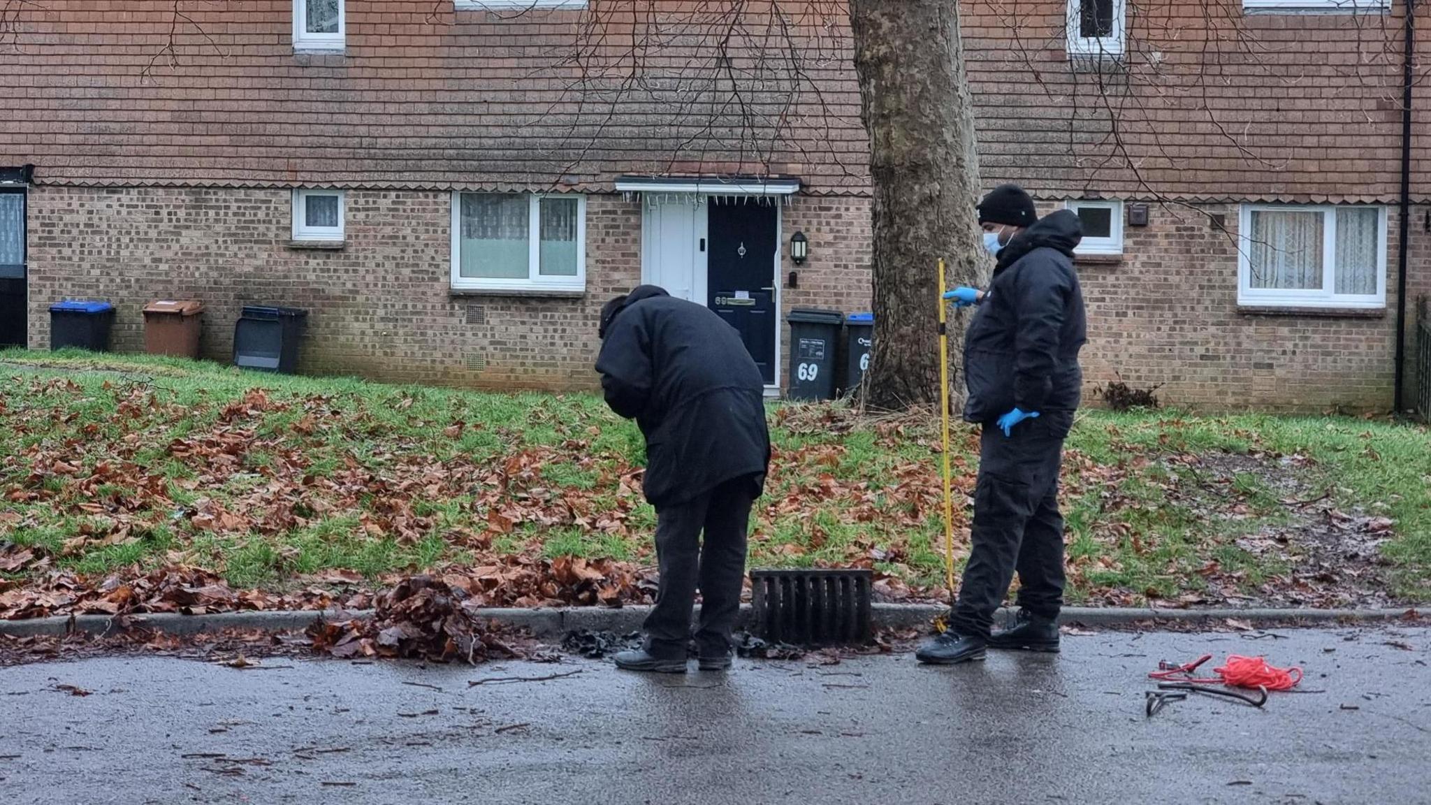 Two police officers dressed in black with masks and blue rubber gloves look into an opened drain by a kerb in a housing estate.