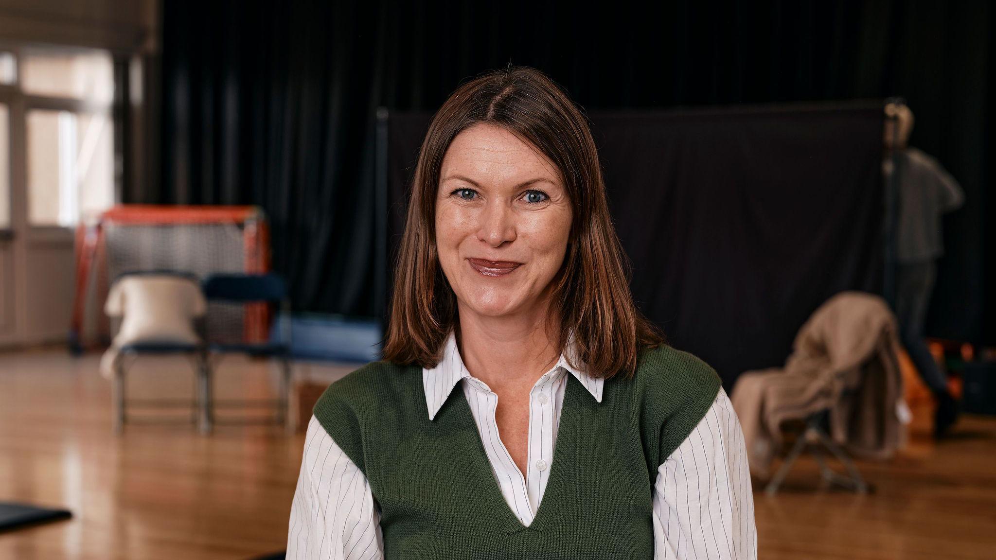 A picture of the charity's CEO. She is smiling wearing brown hair. She is wearing a white striped shirt with a green vest on top.