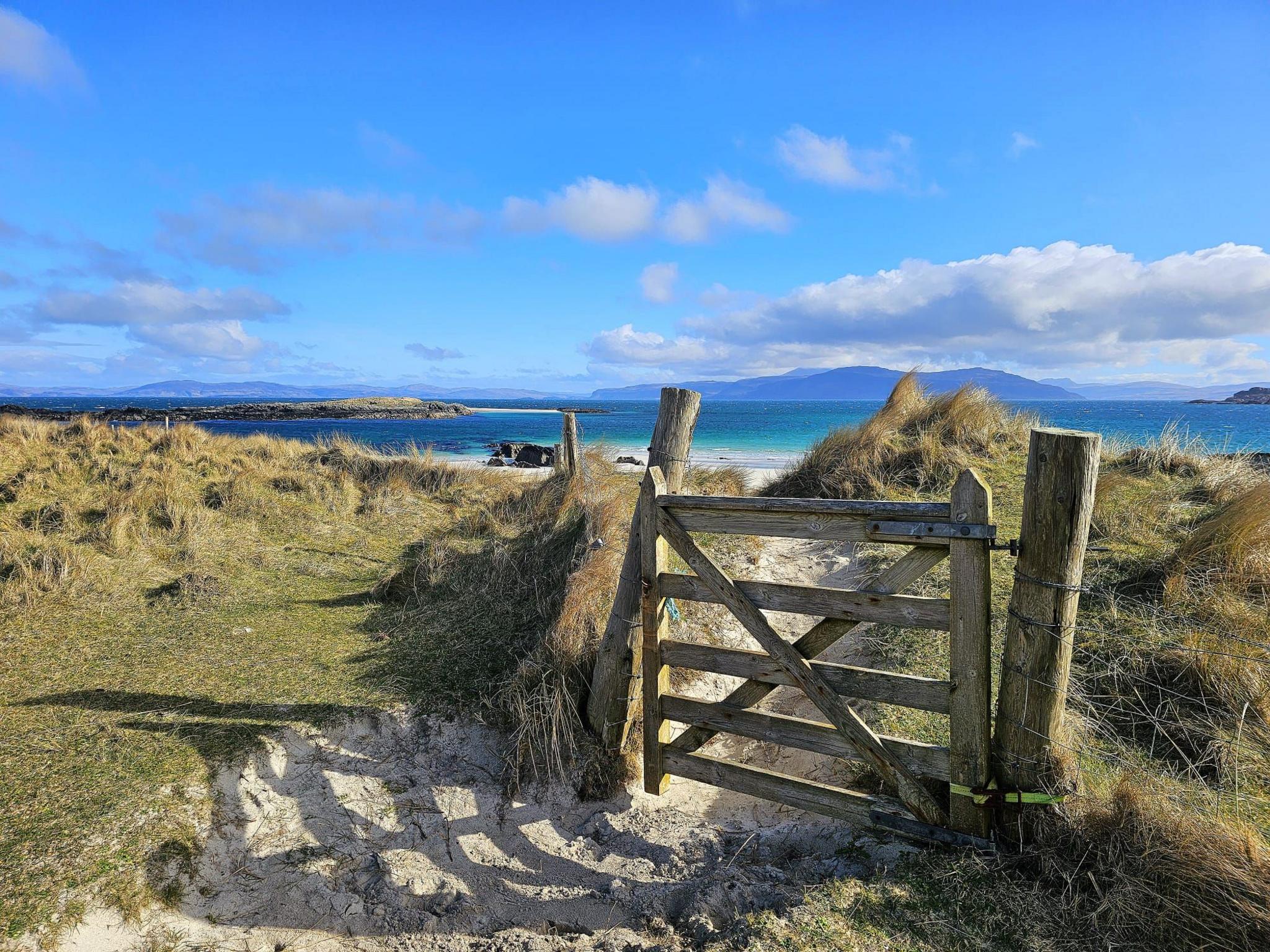 A wooden gate leading down to a beach, with the sun shining over grass the loch beyond it