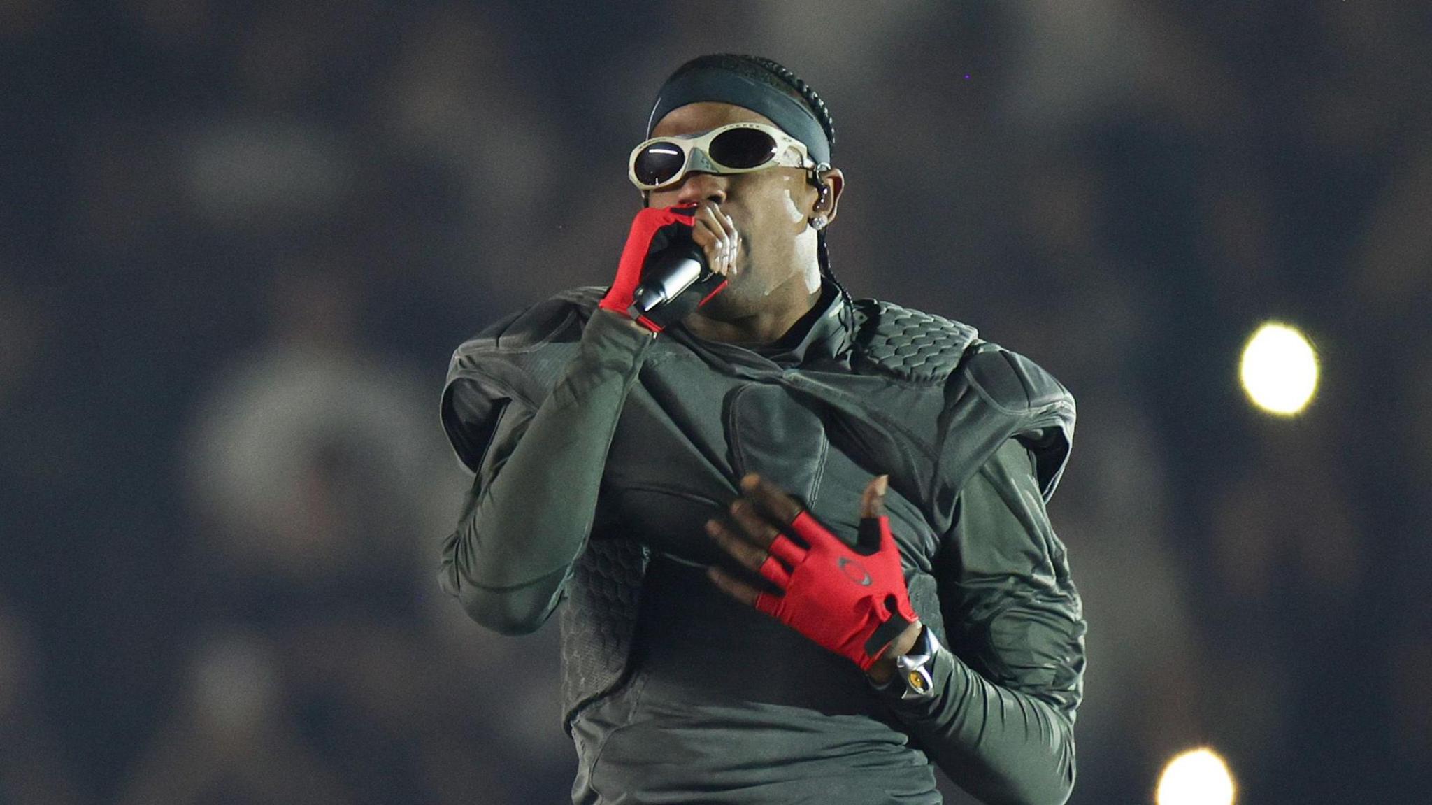 Travis Scott performing on stage. He wears a black outfit that resembles an American football kit, with padded shoulders and red gloves. He wears sunglasses as he sings into a mic, one hand on his chest. 