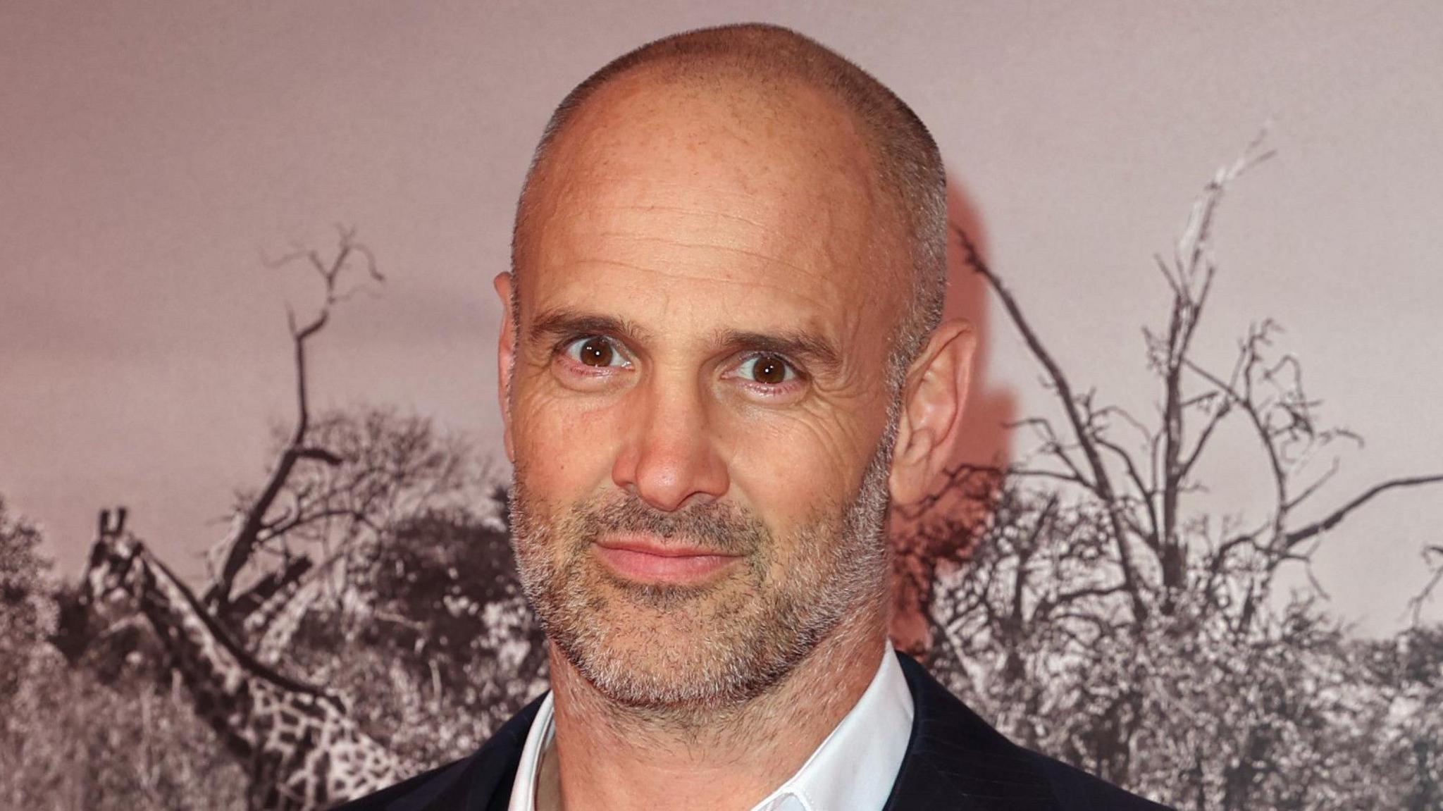 A bald man in a white shirt and pin stripe blazer stands against a wall