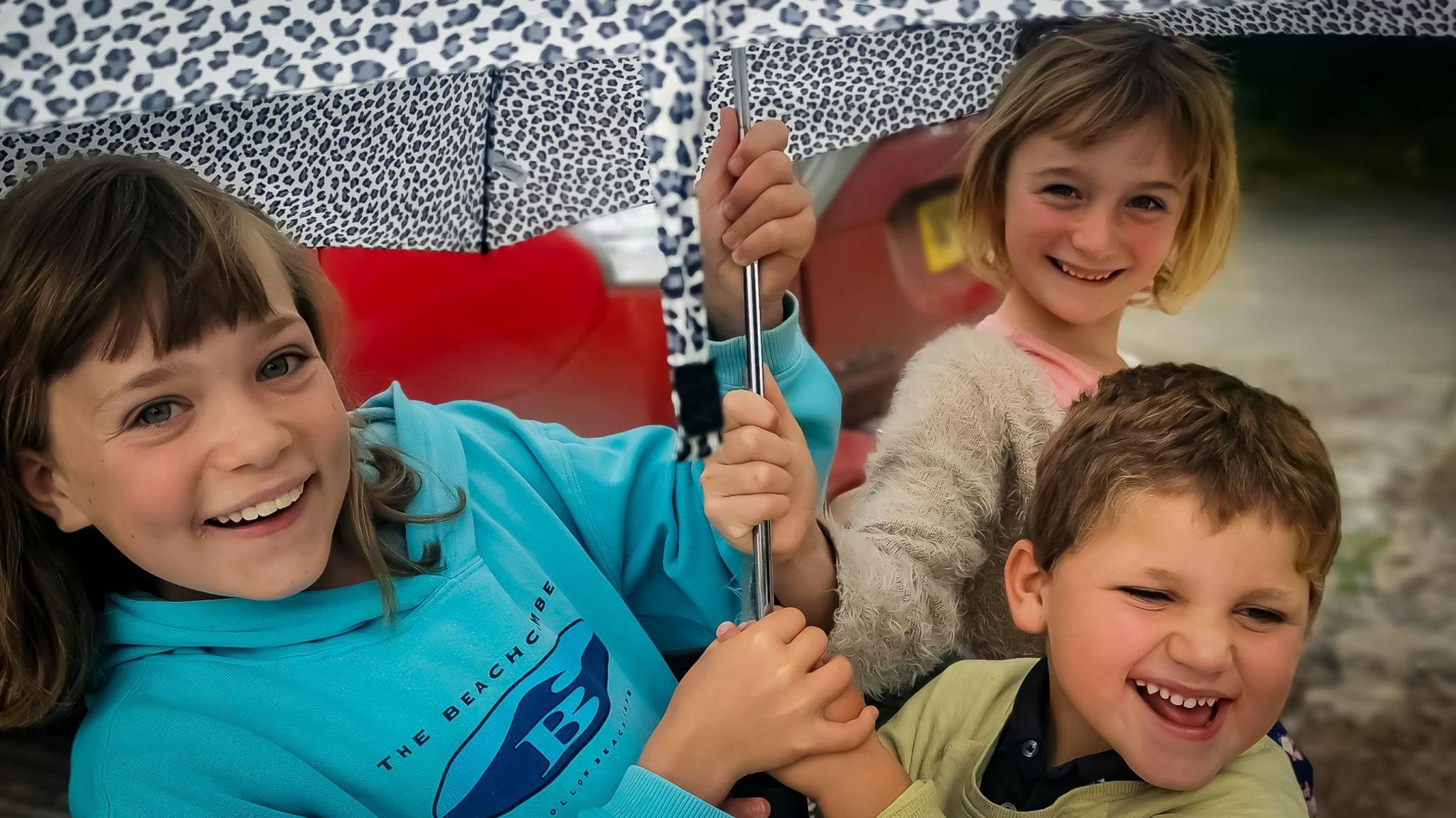 Bethan, Olwen and Ivor have their hands on the stem of an umbrella. They are all smiling. Bethan has shoulder length brown hair and a light blue hoodie on. has a blond bob cut and a fuzzy cardigan. Ivor's hair is short and he has on a yellow sweater, behind him is Olwen smiling with a blonde bob and white furry cardigan on.