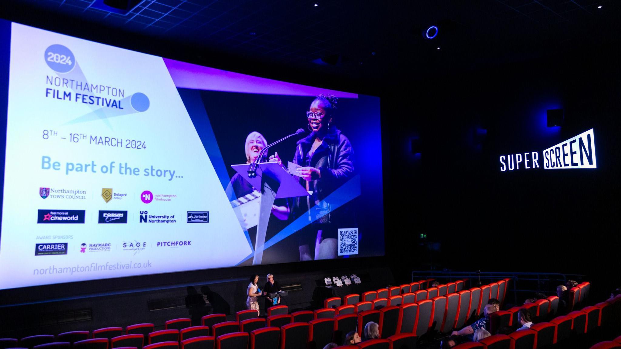 A cinema-like room featuring a large screen highlighting the Northampton Film Festival and rows of red seats. Details of the event and 11 sponsors are listed on the screen. There are red seats in the auditorium. 