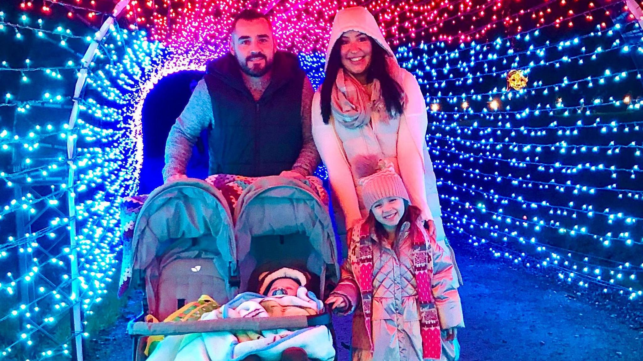 Sophie Jenkins and her husband smiling in a light display, along with Ronnie, one, in a stroller, and Elsie, standing alongside it and in front of her parents