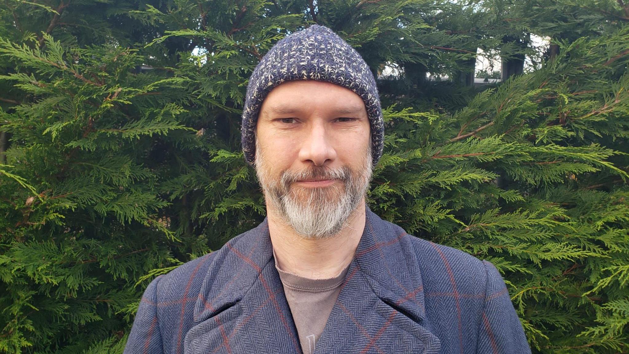 Matthew Pocock with a short white beard standing outside. He is wearing a blue knitted beanie and a blue tweed jacket with a t-shirt.