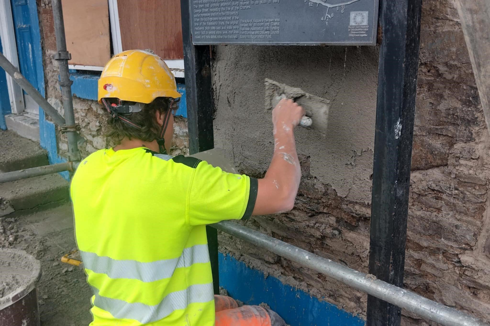 A man in a high-viz top and yellow hard hat is plastering on traditional stone at the town hall