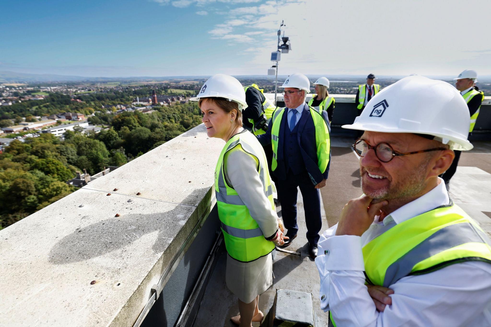 A man and a woman in hi-viz vests and white construction helmets looking over the roof of a building
