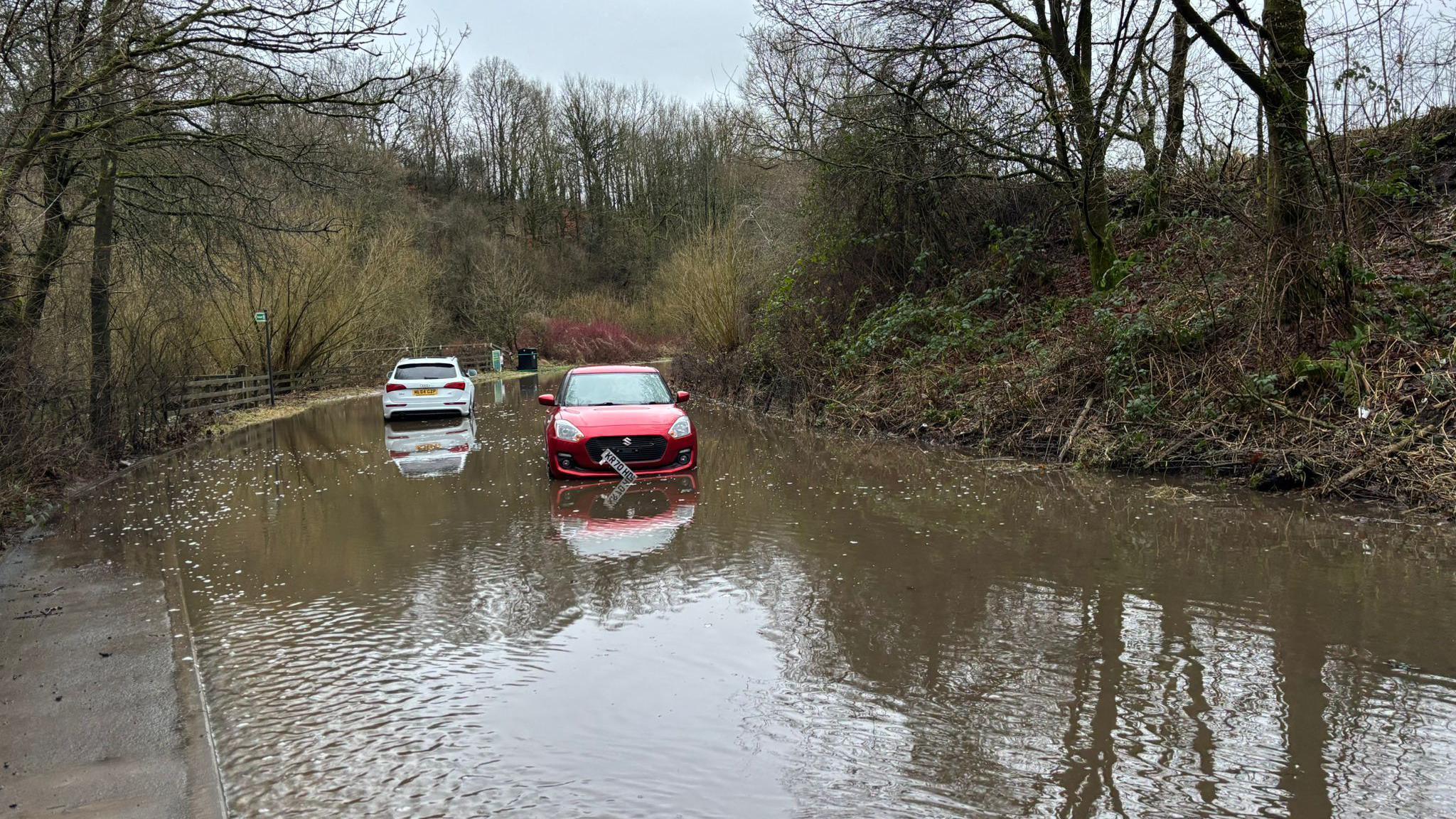 A red and white car stuck in floodwater by a tree-lined bank 