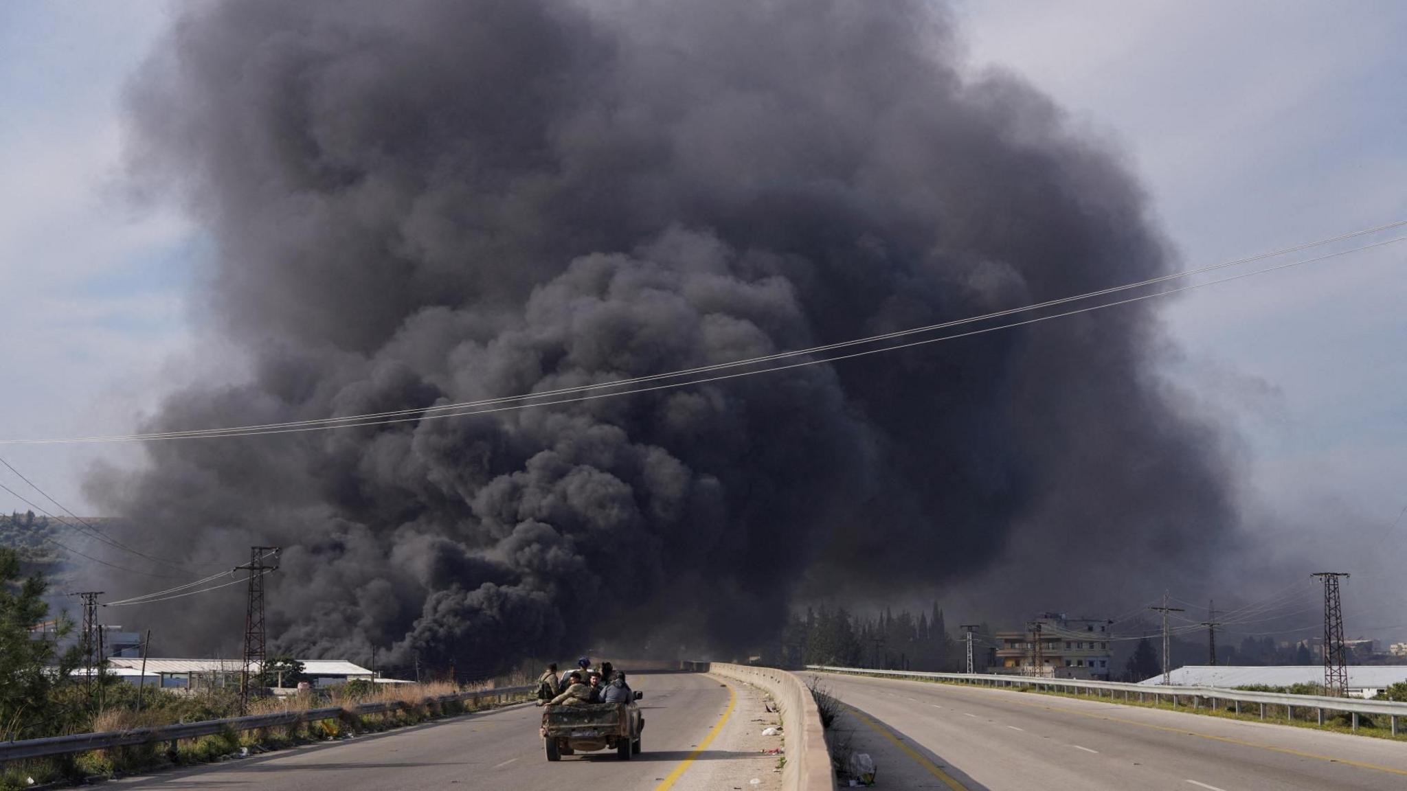 Smoke rises while members of the Syrian security forces ride on a pick-up truck during fighting with insurgents loyal to former president Bashar al-Assad, in Latakia (7 March 2025)