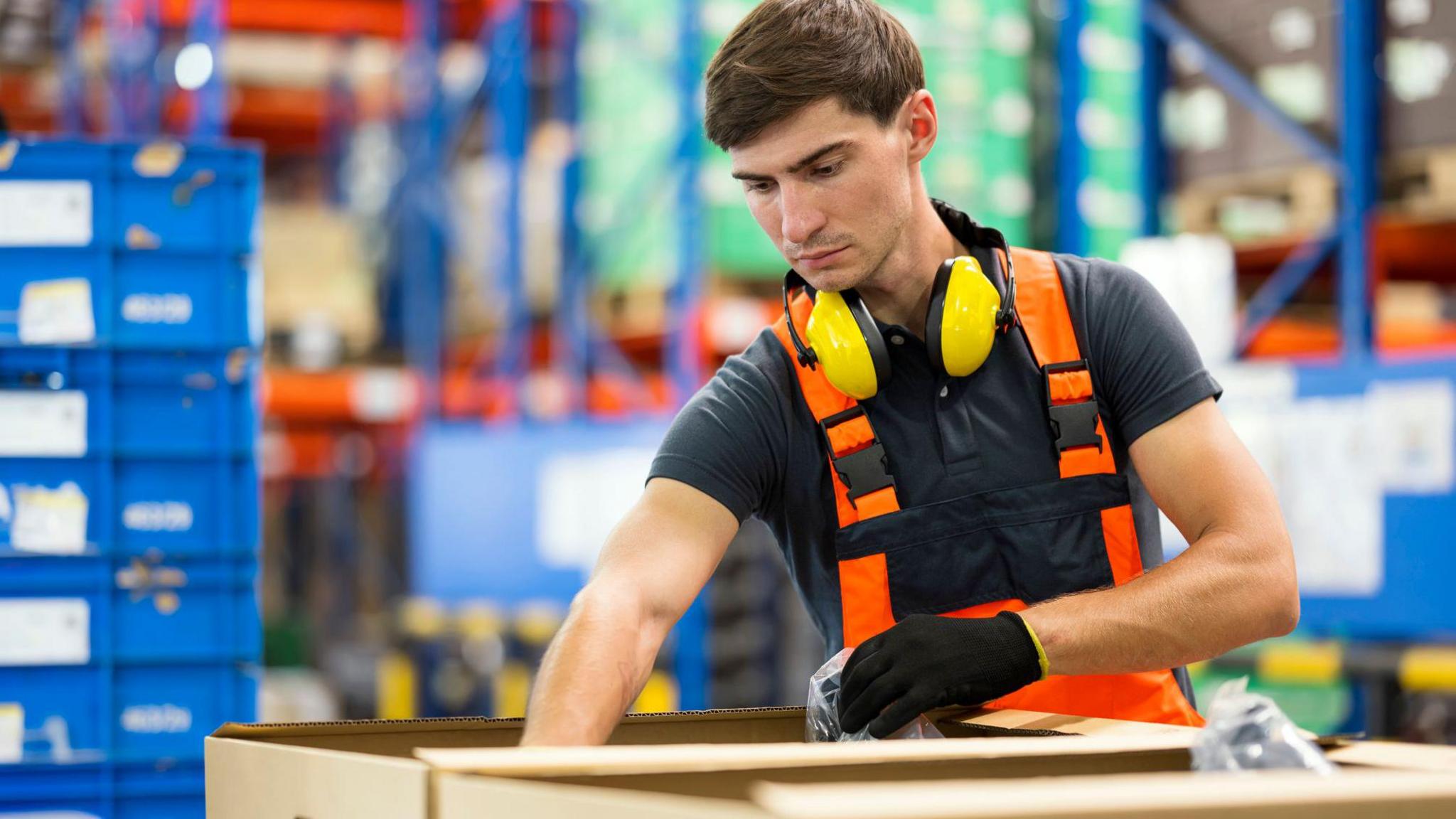 A factory worker wearing an orange work vest, ear defenders around his neck and gloves, puts items in a cardboard box in a warehouse