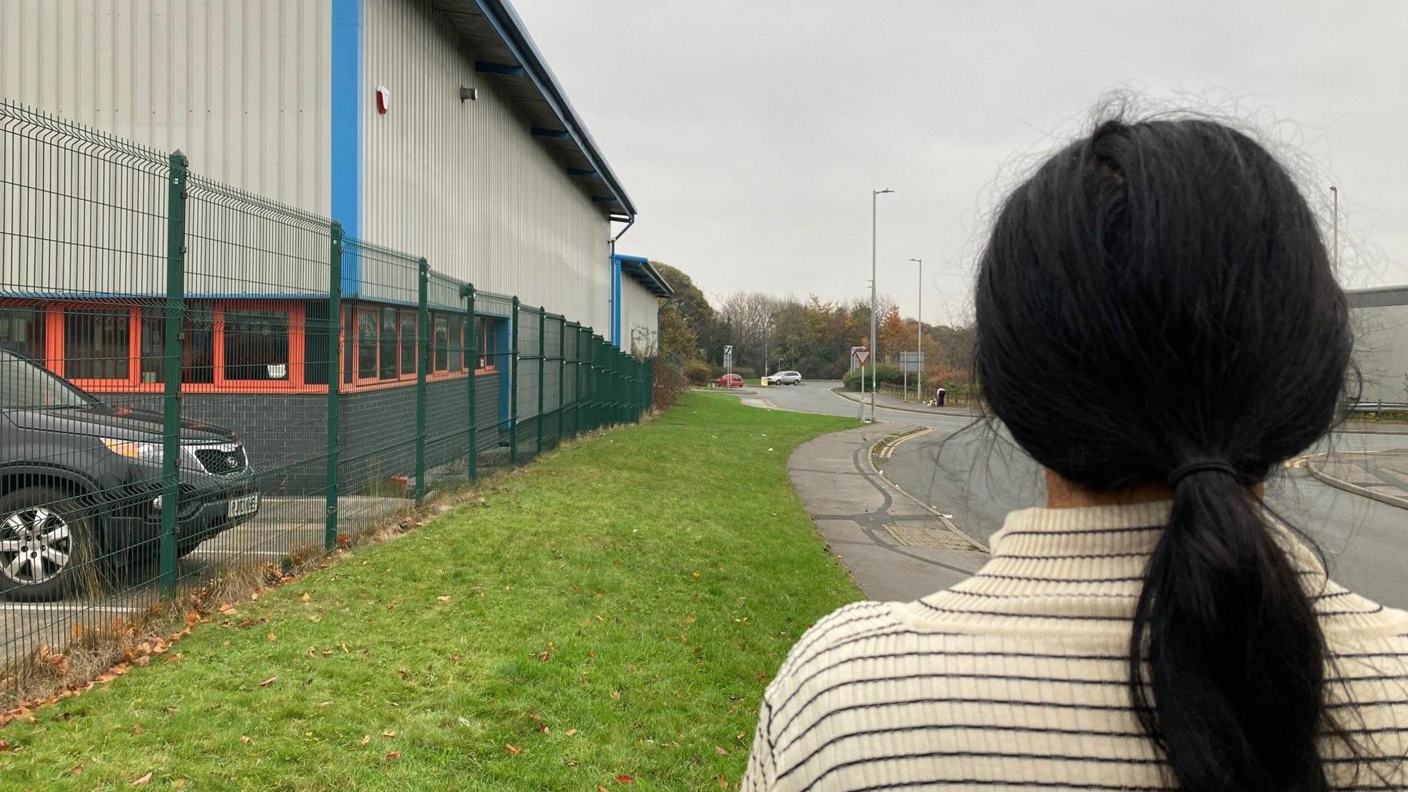 The back of a woman's head with dark black hair in a ponytail and a black and cream striped top. A green grass verge stretches out in front of her by the side of warehouse building.