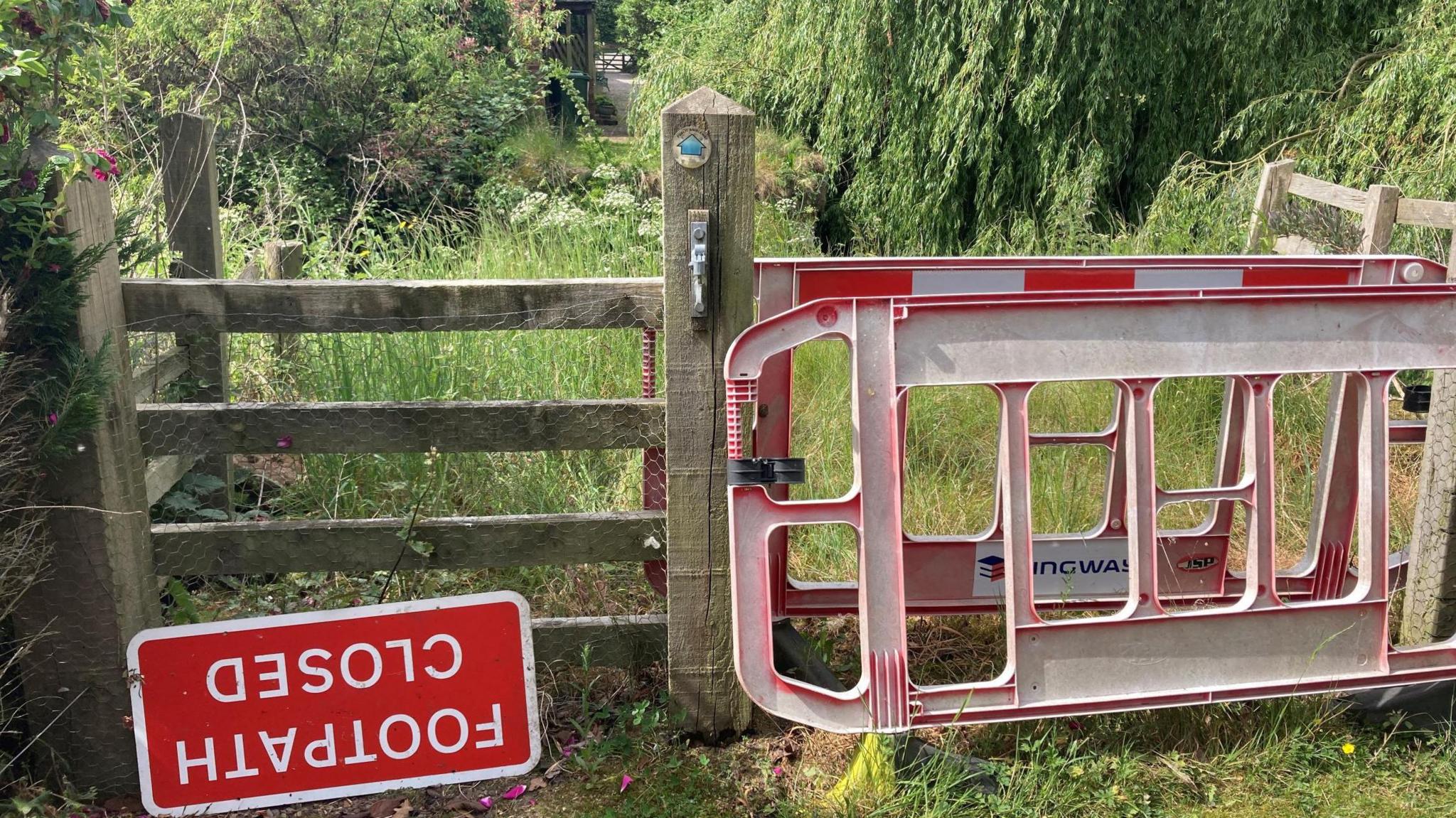 Stile with an upside down  'footpath closed' sign in front.  There are red plastic barriers leaning alongside the stile. Over the stile is undergrowth and a house and path in the distance.  