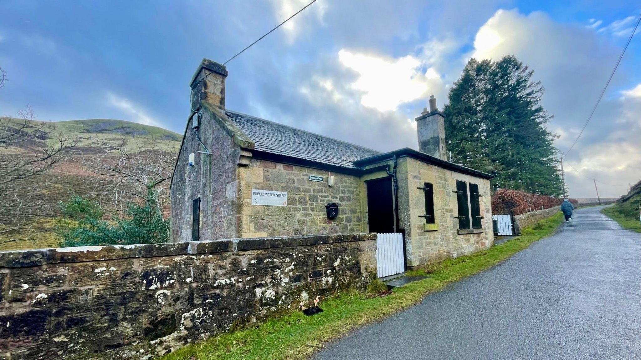 A paved road passes through the picture beside Loganlea Cottage. It is stone built and has stone wall. Some trees and a hill are behind it.