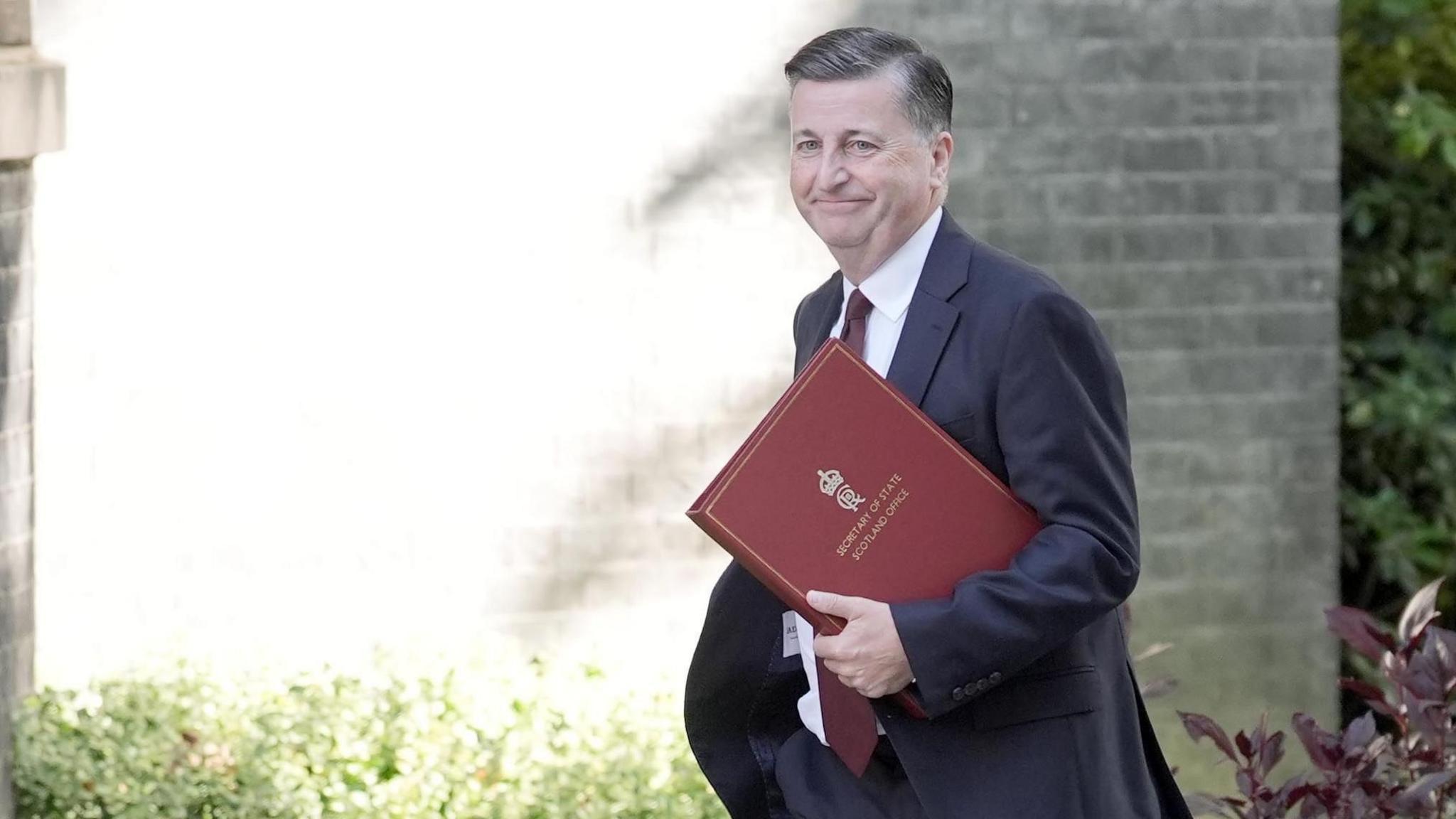Douglas Alexander smiles while walking into Downing Street. He is wearing a blue suit, white tie and a maroon tie and is holding a maroon folder with Secretary of State Scotland Office written on it.