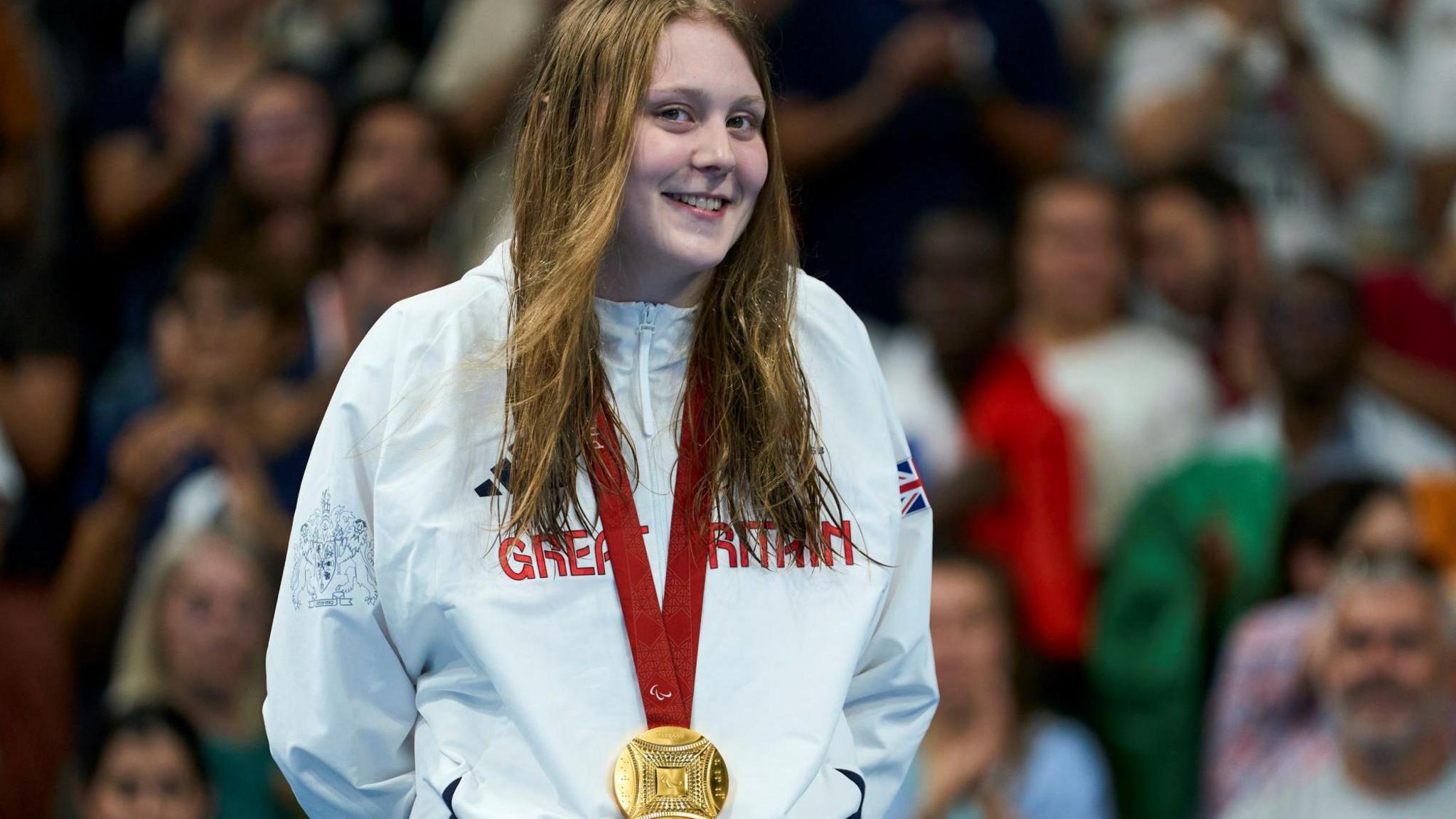 Poppy Maskill on the podium after winning the gold medal in the Women's 100m Butterfly She is wearing a gold medal and has a Great Britain tracksuit top on. She has long hair and is smiling at the camera. 