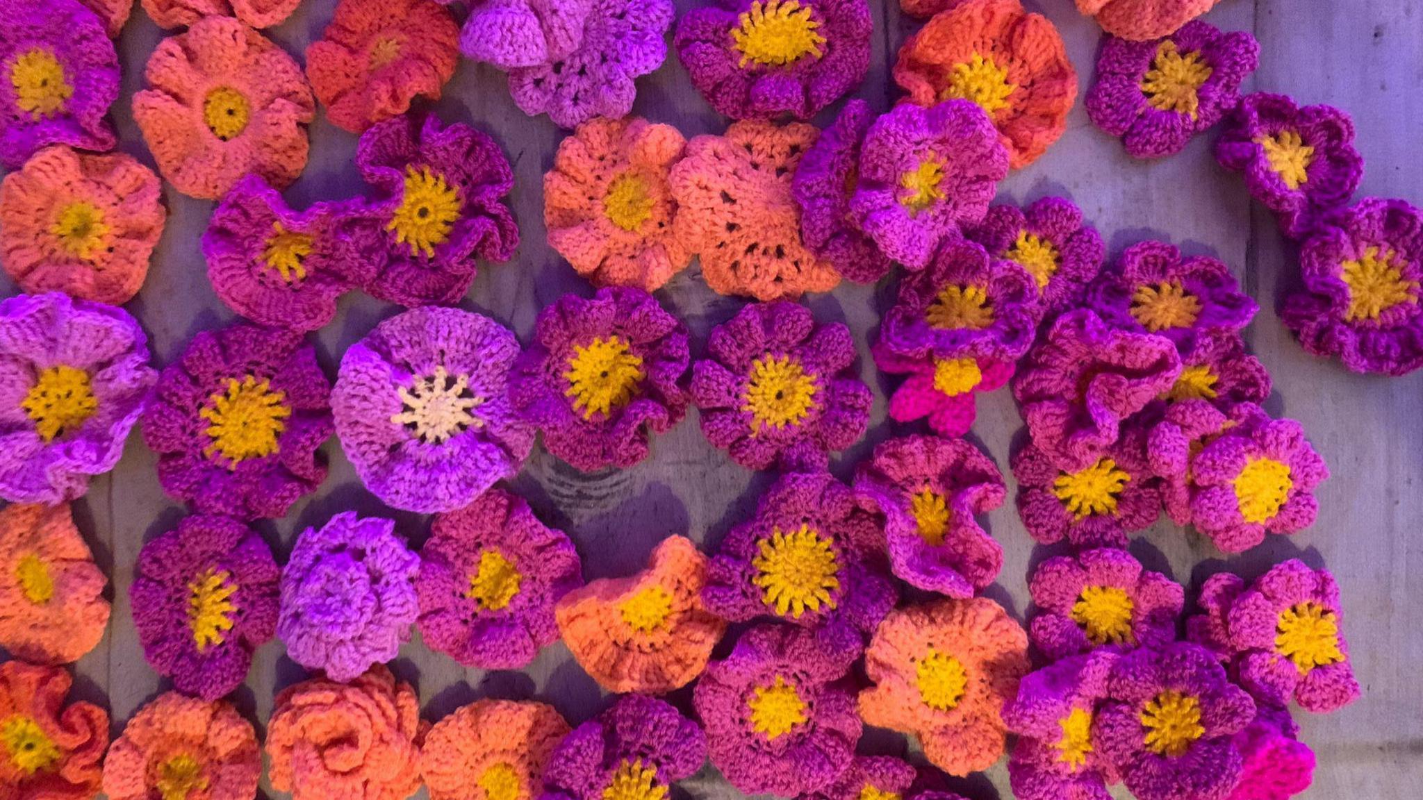 A table is covered in palm-size crochet flowers. They are purple, pink and orange. 