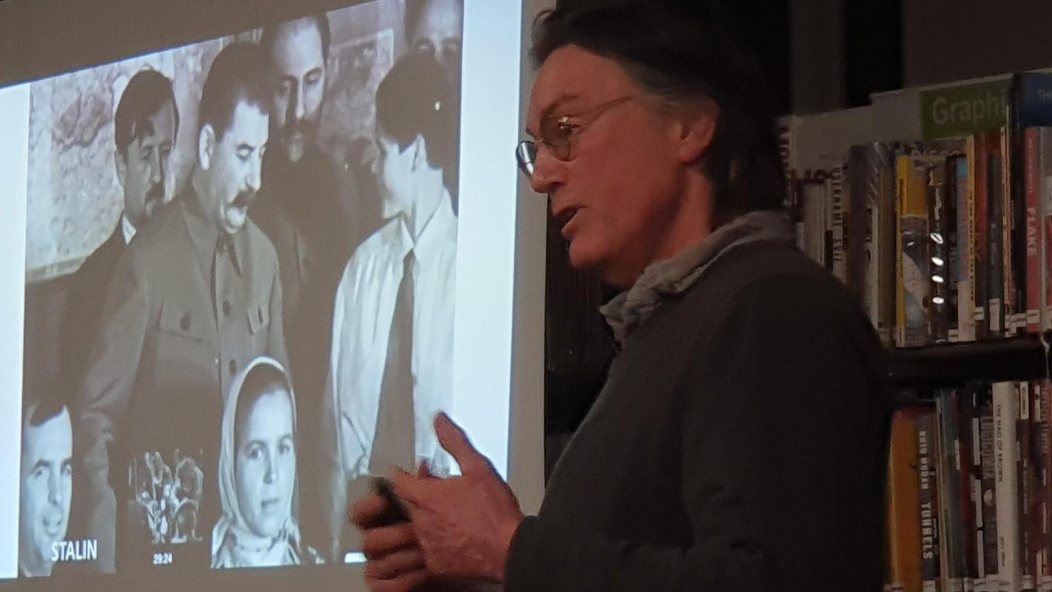 Philip Colley giving an animated lecture on his great uncle. His hands are clasped in front of his chest. He has a grey jumper over a grey shirt. He has rounded glasses and short dark hair. To his right is a screen showing an image of Stalin, standing among a group of people. Behind him is a shelf full of books.