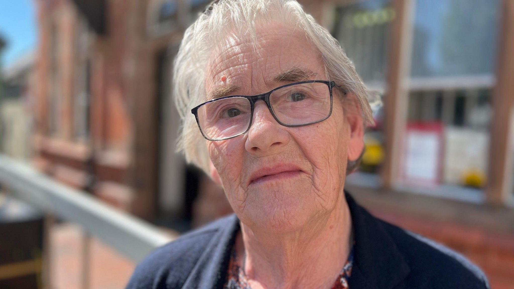 An 86-year-old woman with grey hair wears dark glasses and a dark top is looking straight at the camera.