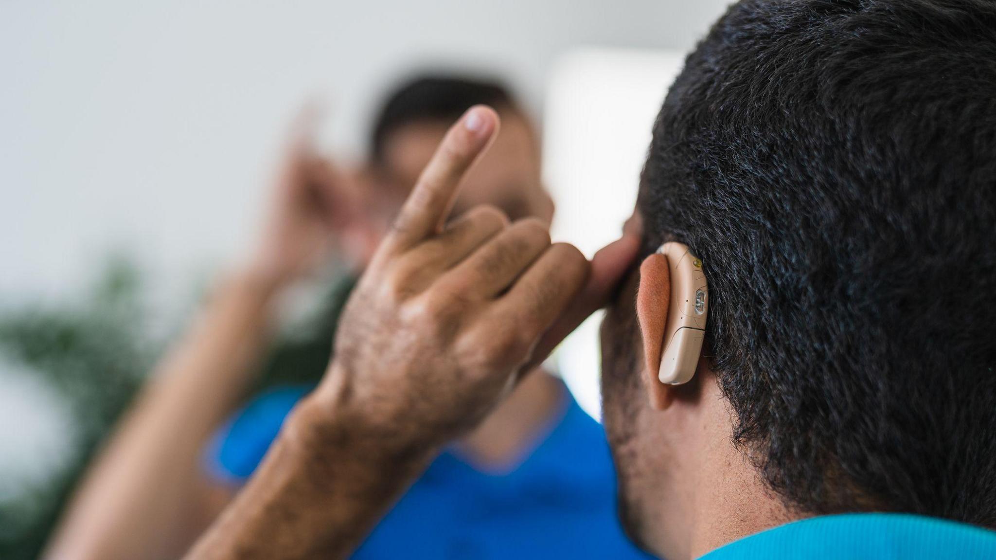 A man is facing away from the camera. He has a hearing aid on his ear, and his hand is up to his head. He is signing.