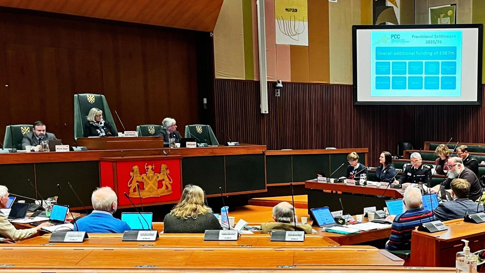 Alison Hernandez, James Vaughan and various councillors sit in a council chamber for the Police and Crime Panel.