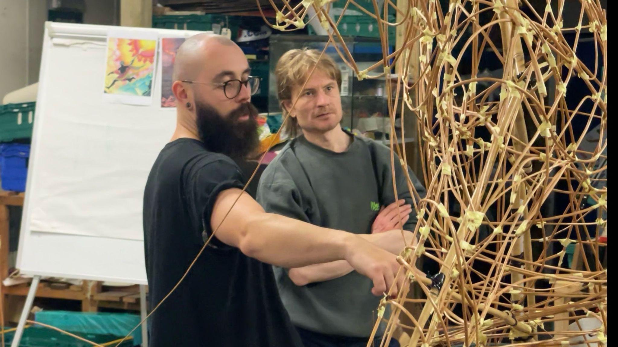 Two men stand beside a large complex structure made from willow canes and masking tape. The man on the left has a bald head, bushy beard, glasses and jeans. He is pointing at the structure whilst speaking. The man on the right has a grey jumper rolled up to his sleeves and long fair hair. He has his arms crossed and is observing the structure. A flip chart can been seen in the background with a picture of a Greek god attached. They are inside a large workshop with numerous boxes of tools and supplies on shelves.