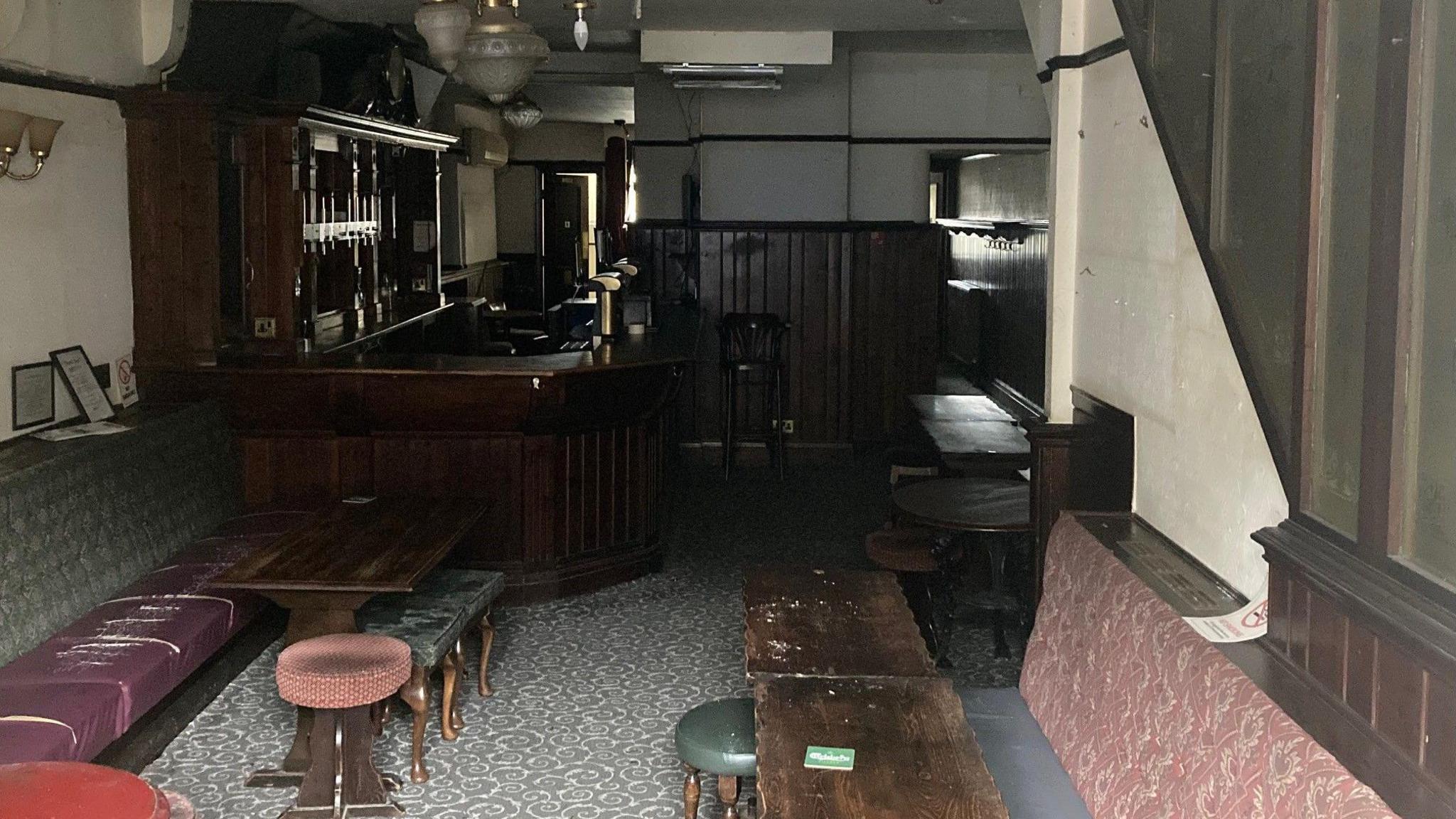 Fabric and leather topped bar stools next to dark wooden tables in the foreground with a dark wood bar to the right-hand side. You can see the outlines of where photographs were once hanging on the yellow-white stained walls.