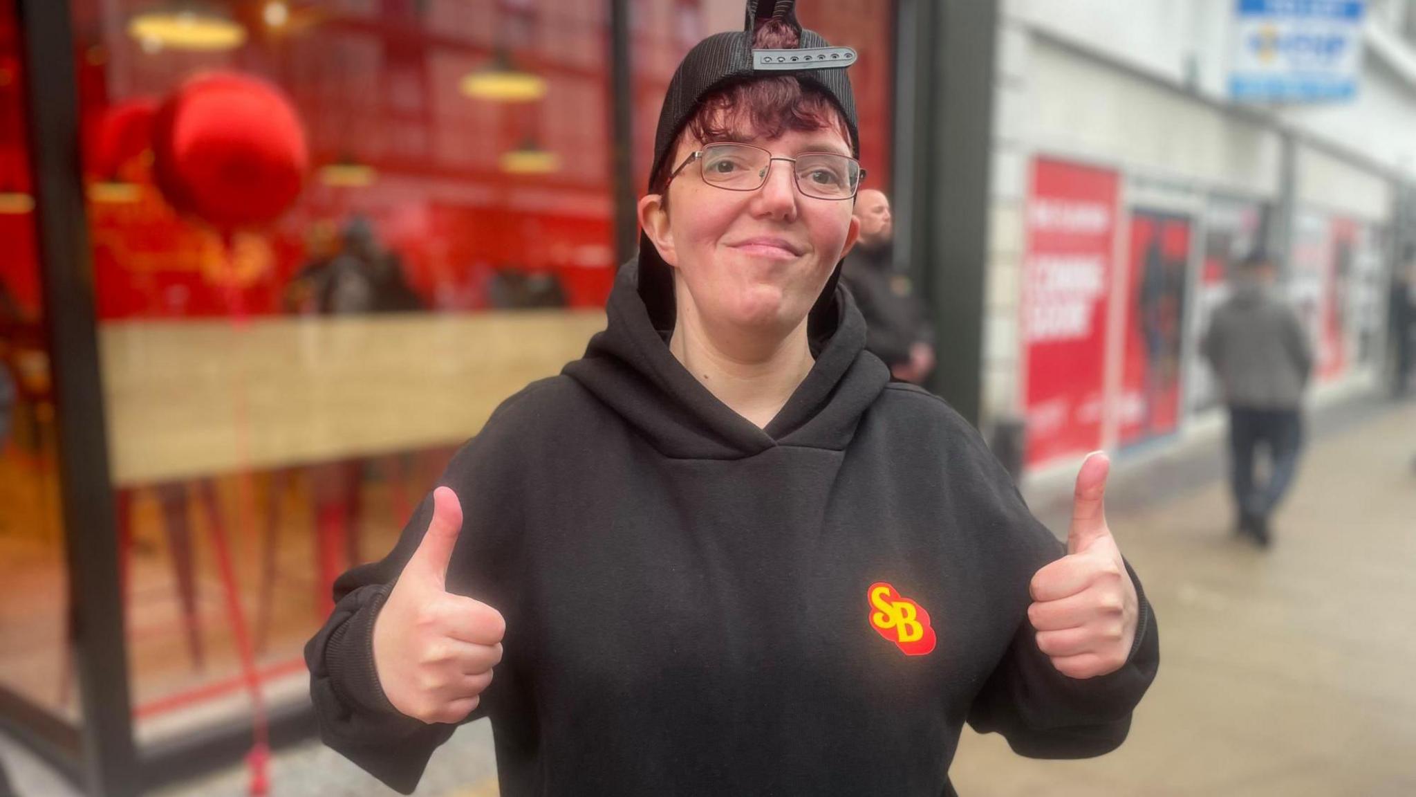 A woman with a black hoodie and black baseball cap turned backwards, smiles at the camera holding up two thumbs