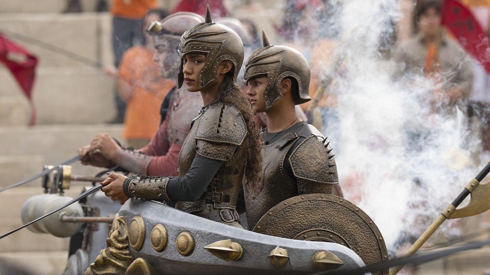 Dior Goodjohn holds reigns whilst stood in a bronze studded chariot. She wears bronze armour and a helmet with a spike in it. 