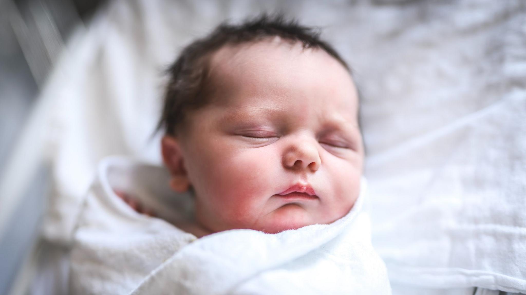 A newborn child, swaddled and lying in a Hospital Bed. The baby has alot of dark hair 